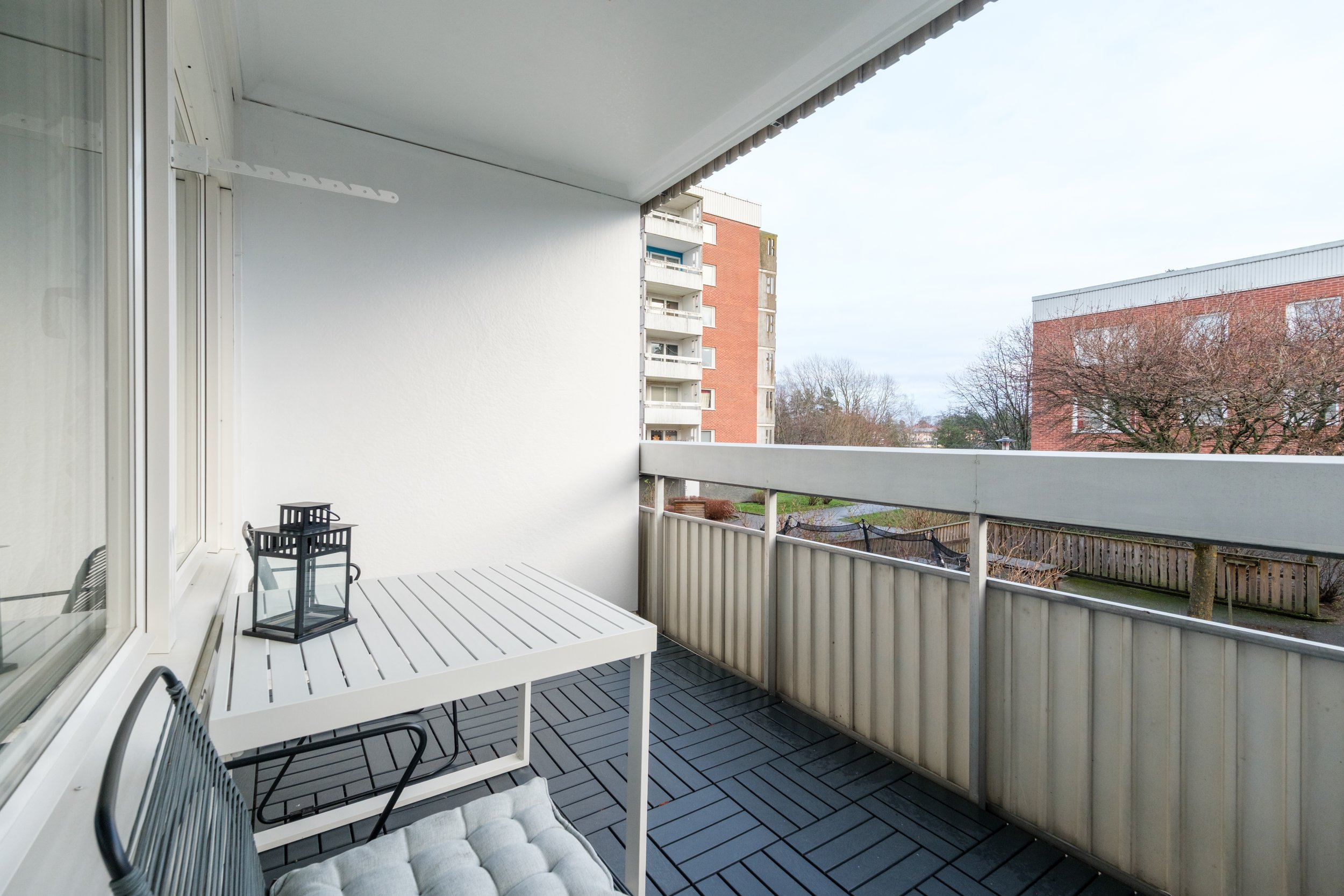 View of a balcony with a white table, black lantern, and chairs, overlooking neighboring apartment buildings and trees.