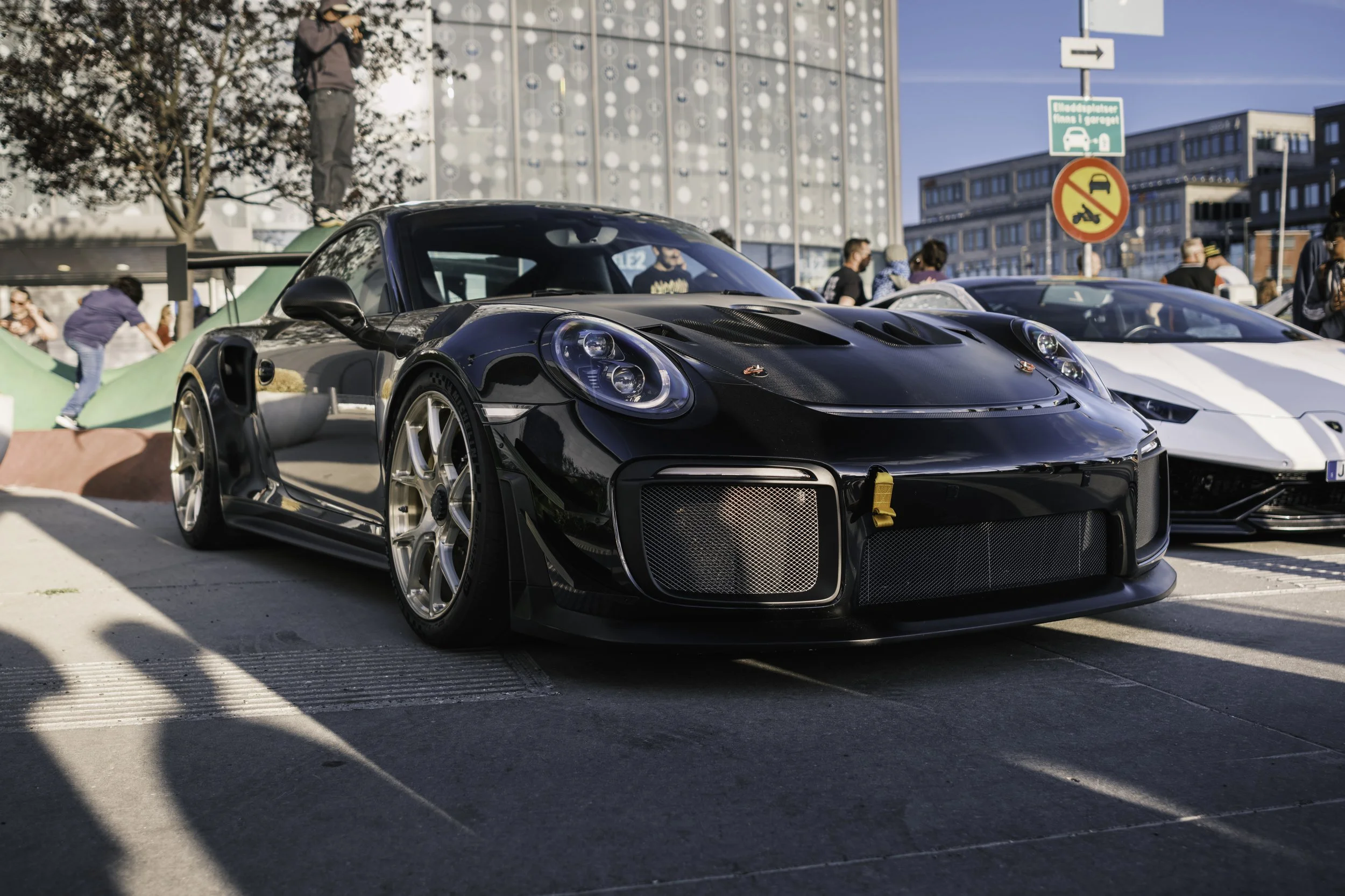 A black Porsche race car parked on a city street during an outdoor car event, with several people and other cars in the background.