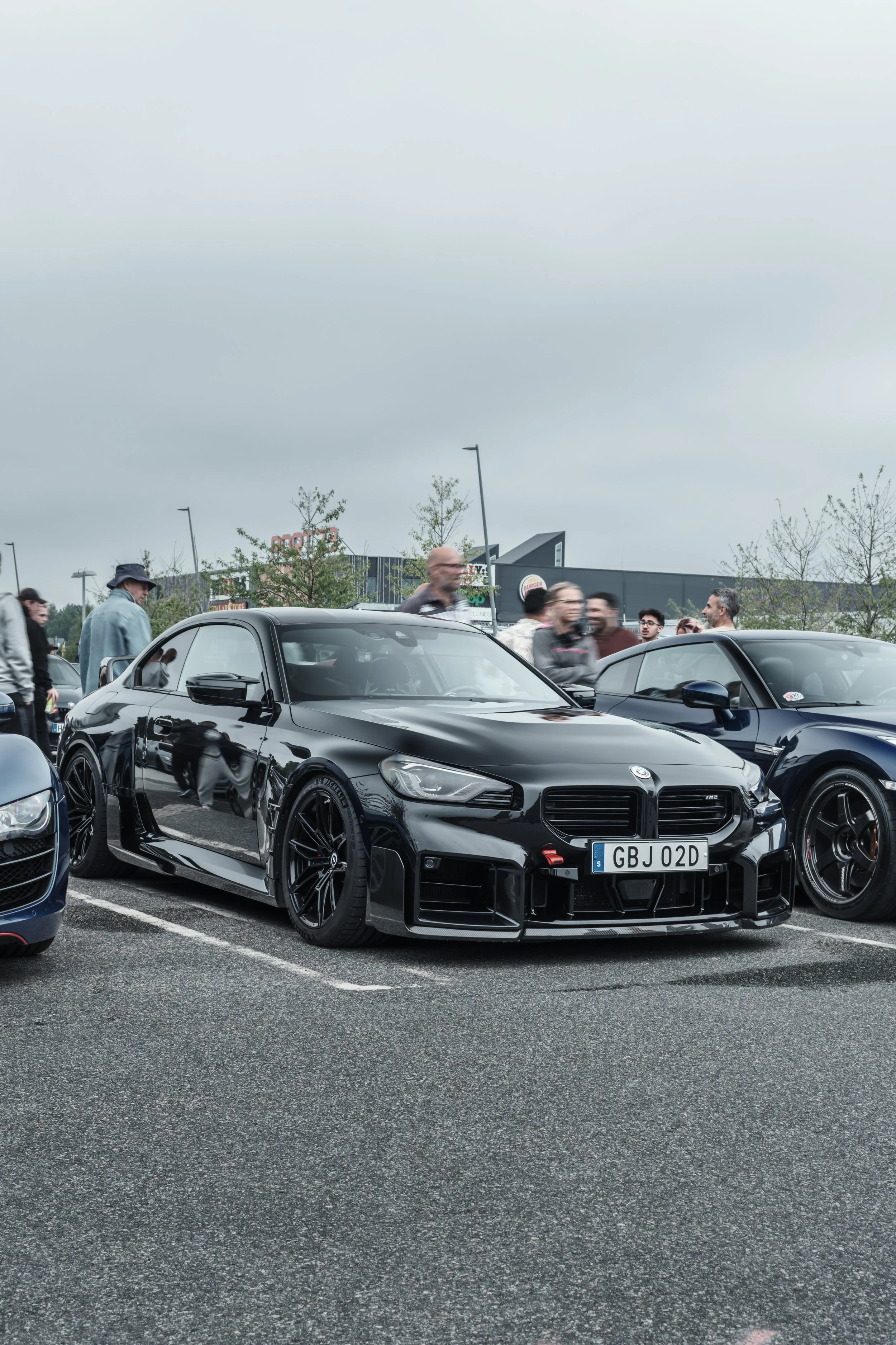 A black high-performance car with a custom front bumper and low stance parked in a lot, with people and other cars visible in the background on a cloudy day.