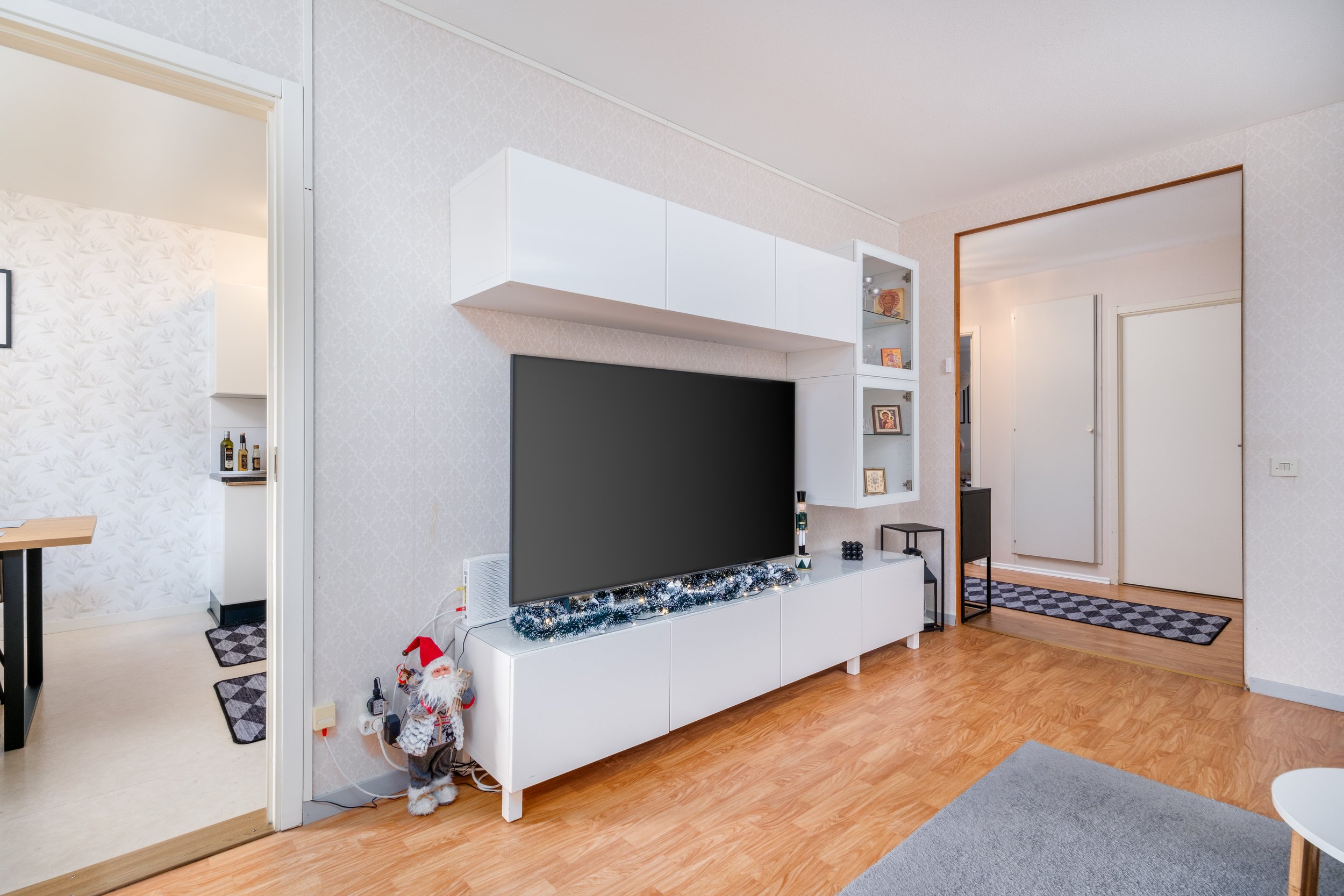 Living room with white storage unit, decorated for Christmas, with a large flat-screen TV, a Santa Claus figure, and a small side table. The room has light-colored walls, wood flooring, and a doorway leading to another room.