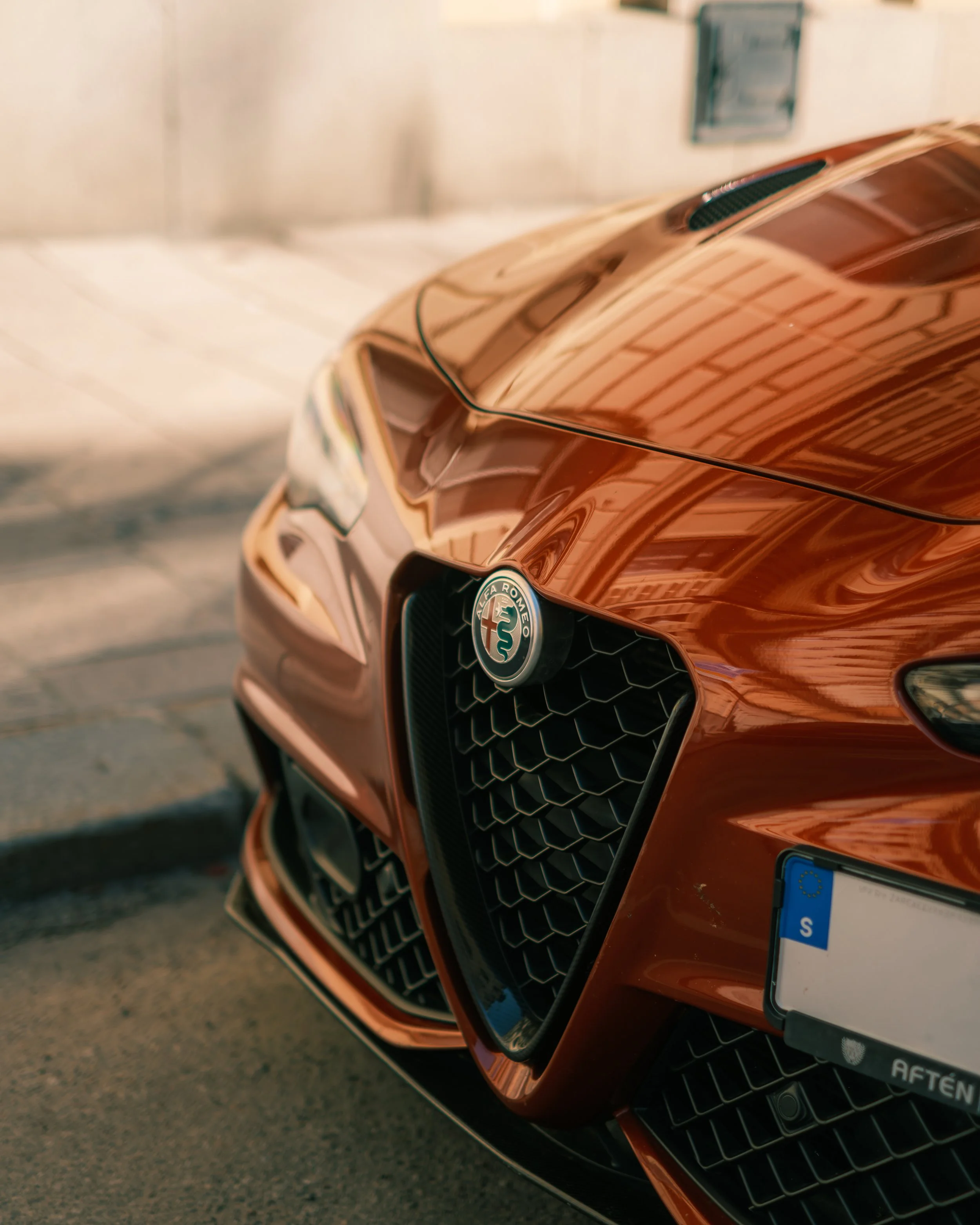 Close-up of the front of a metallic orange Alfa Romeo car, showing the grille with Alfa Romeo emblem and a partially visible European license plate.