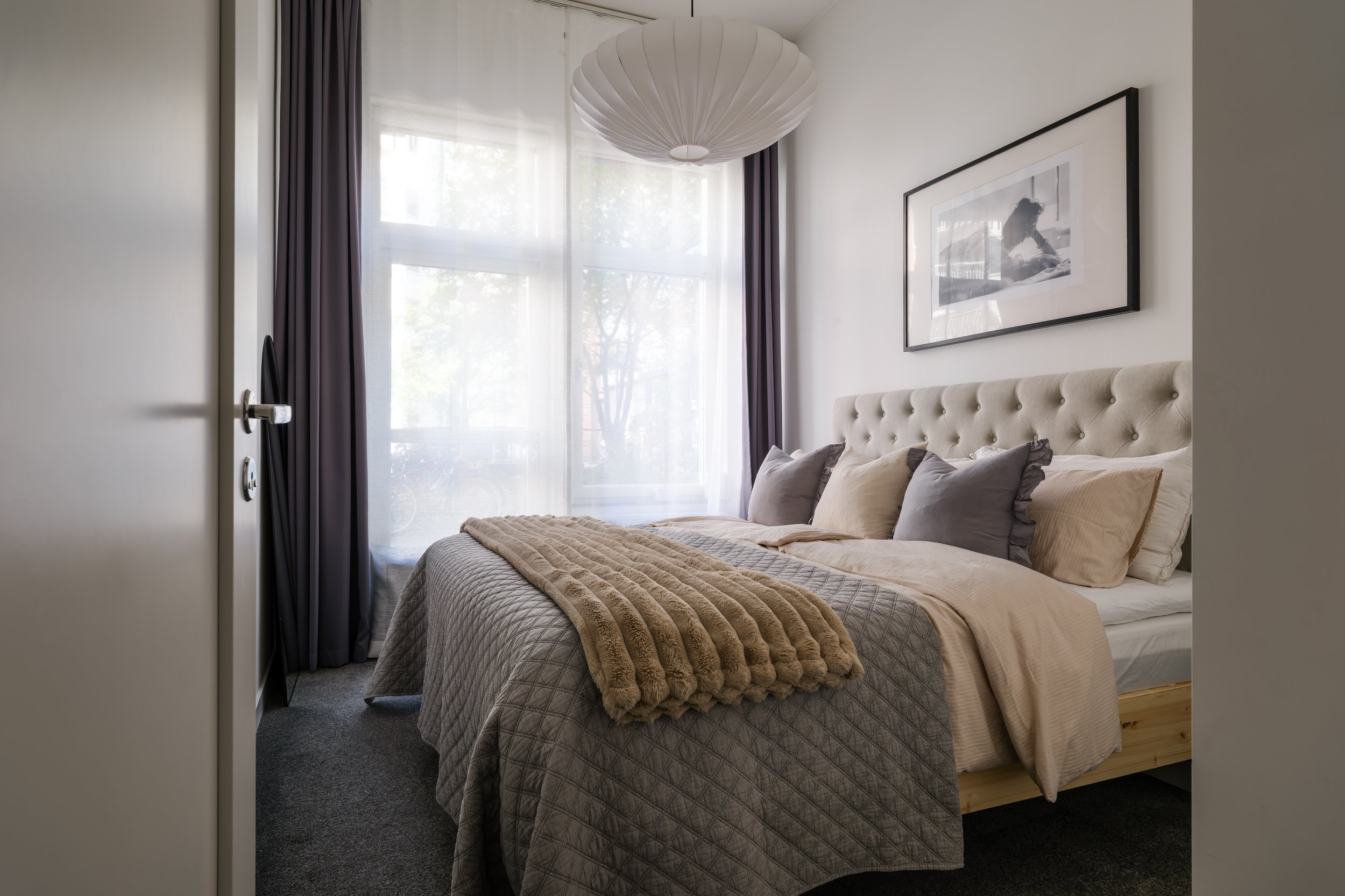 Interior photography of a Scandinavian bedroom with tufted headboard and layered bedding, soft daylight through sheer curtains — Stockholm apartment.