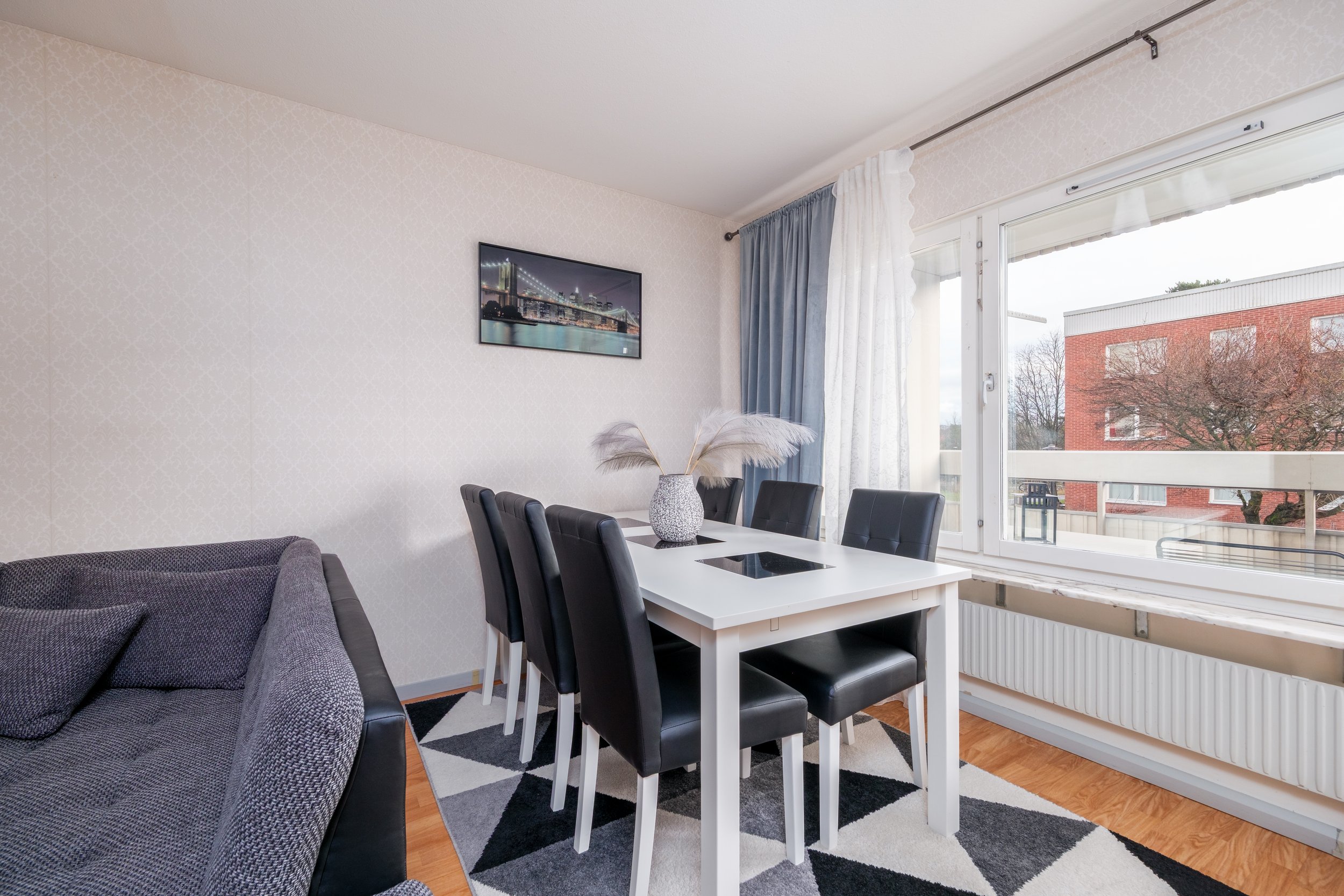 Dining room with a white table, black chairs, a vase with pampas grass, a window with blue and white curtains, and a framed cityscape photo on the wall.