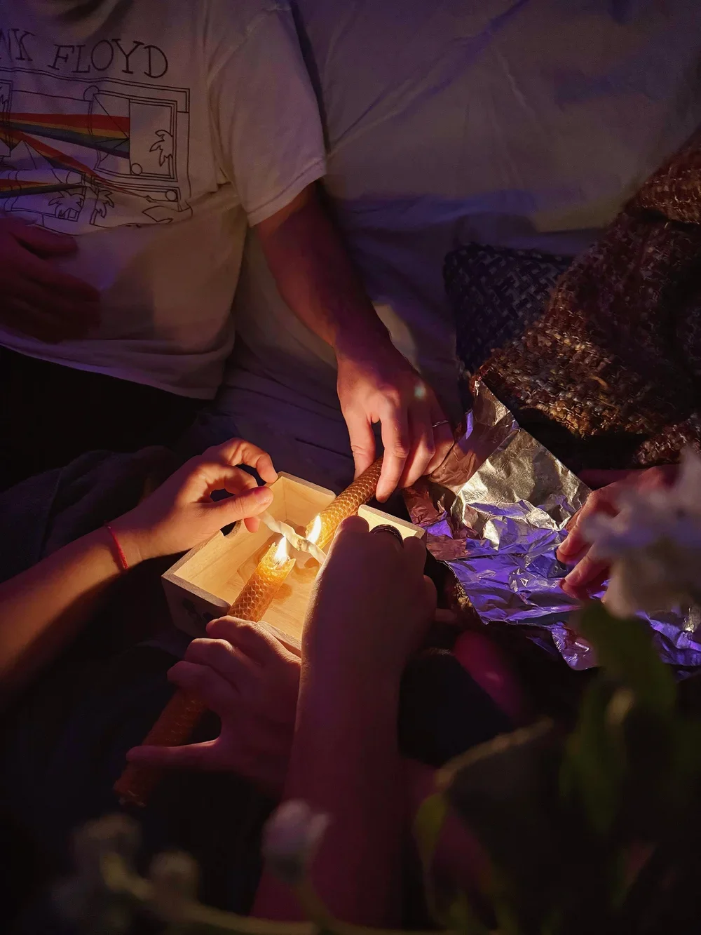 Two people are lighting a candle with a matchstick inside a small wooden box surrounded by foil and fabric. Delayed umbilical cord burning, cutting ceremony right after birth. The scene is dimly lit, creating a warm glow from the candle.