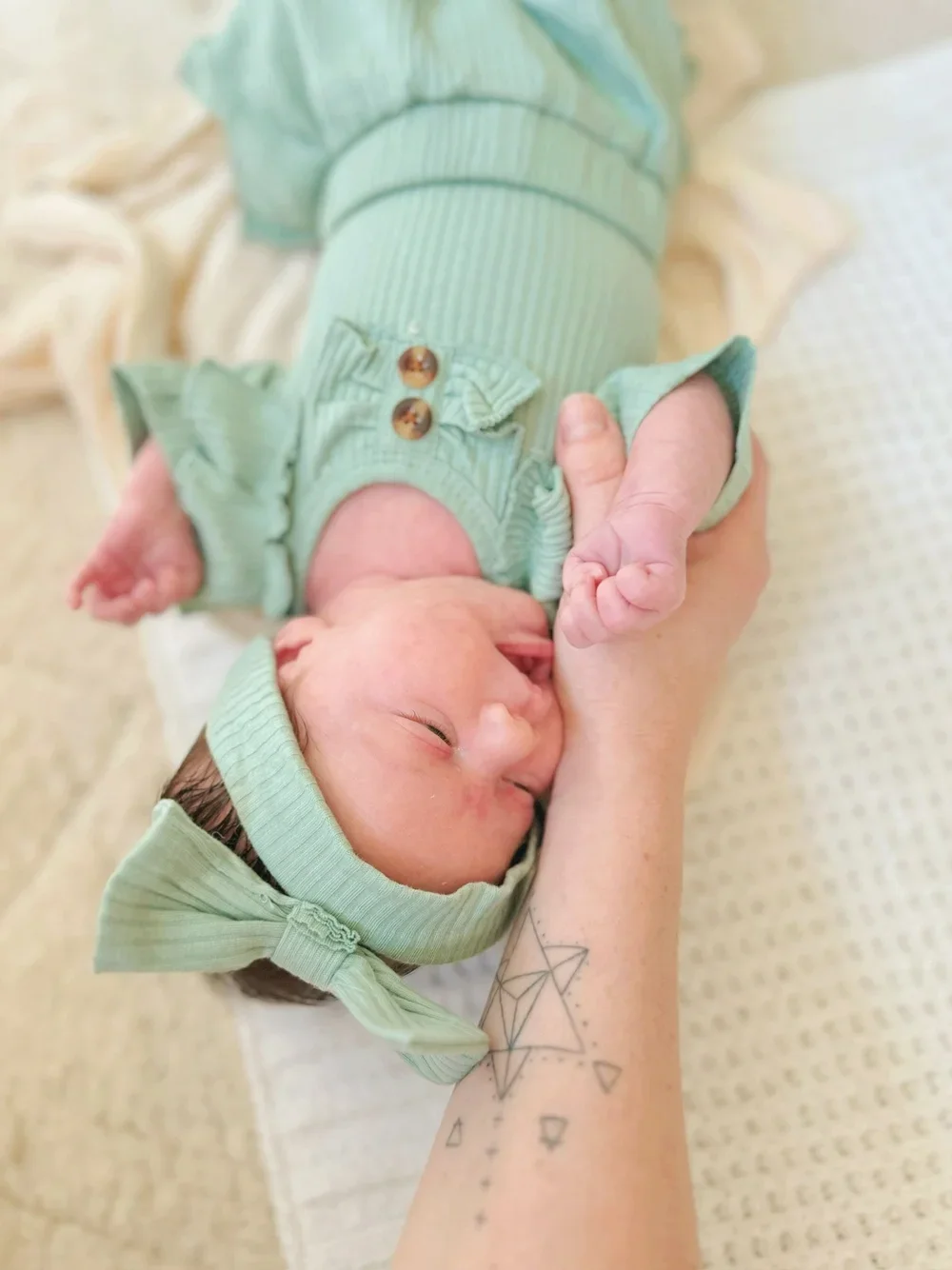 A newborn baby wearing a green outfit and matching headband, lying on a person's arm. The person has a geometric tattoo on their forearm.