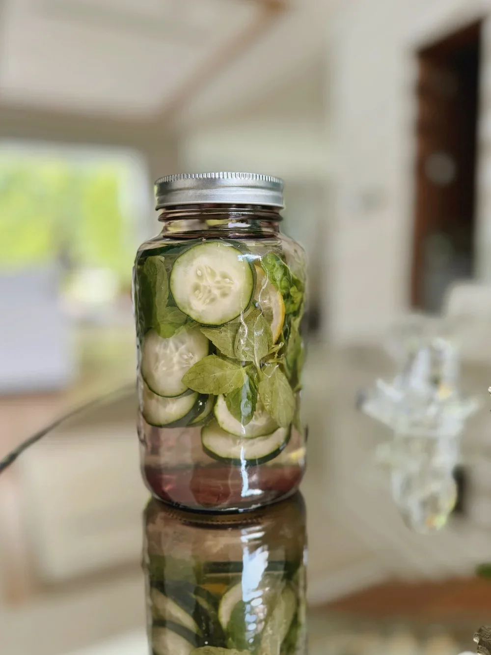 A glass jar filled with sliced cucumbers, basil leaves, and water, placed on a reflective surface indoors.