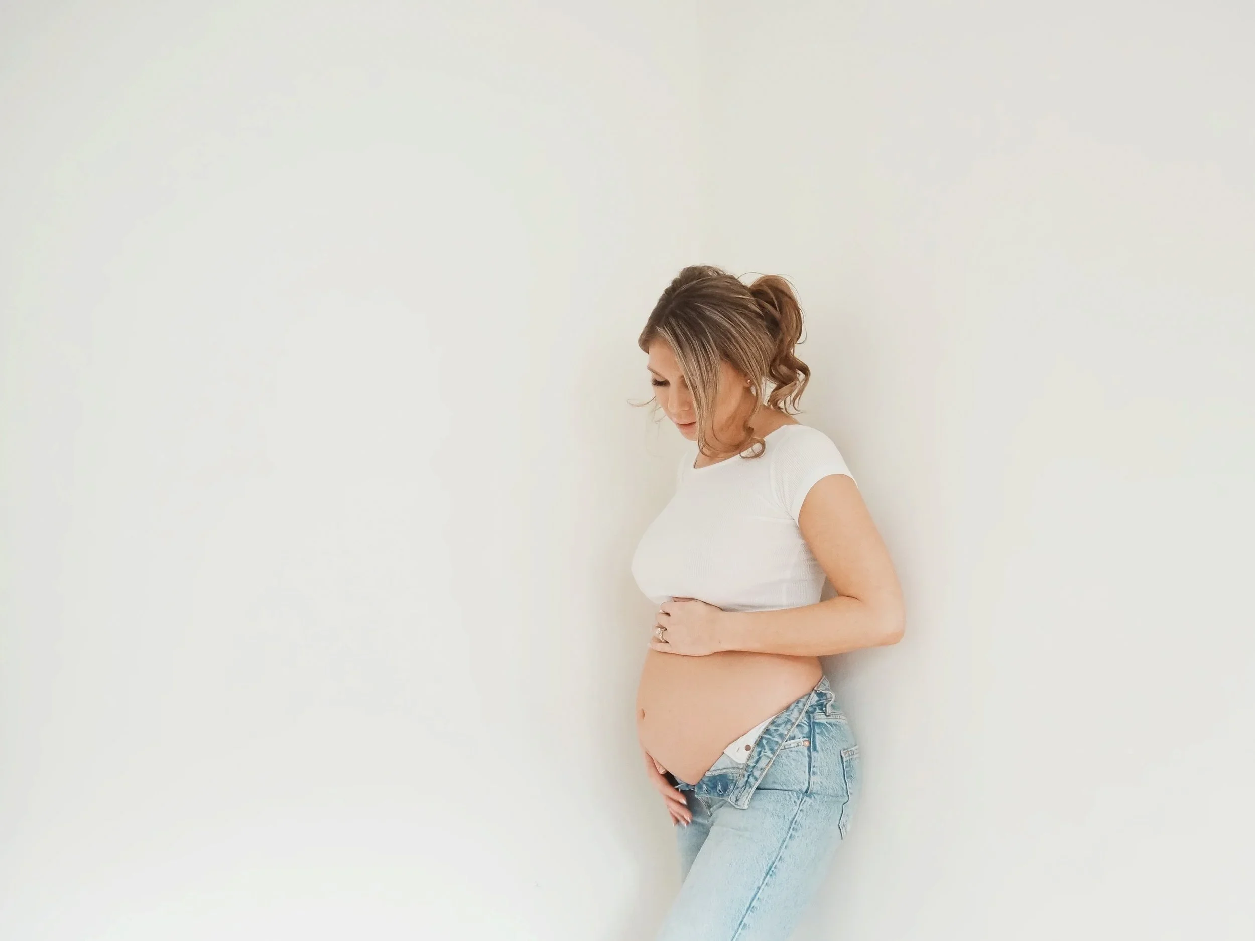 Pregnant woman standing against a white wall, wearing a white t-shirt and light blue jeans, gently cradling her belly and looking down.