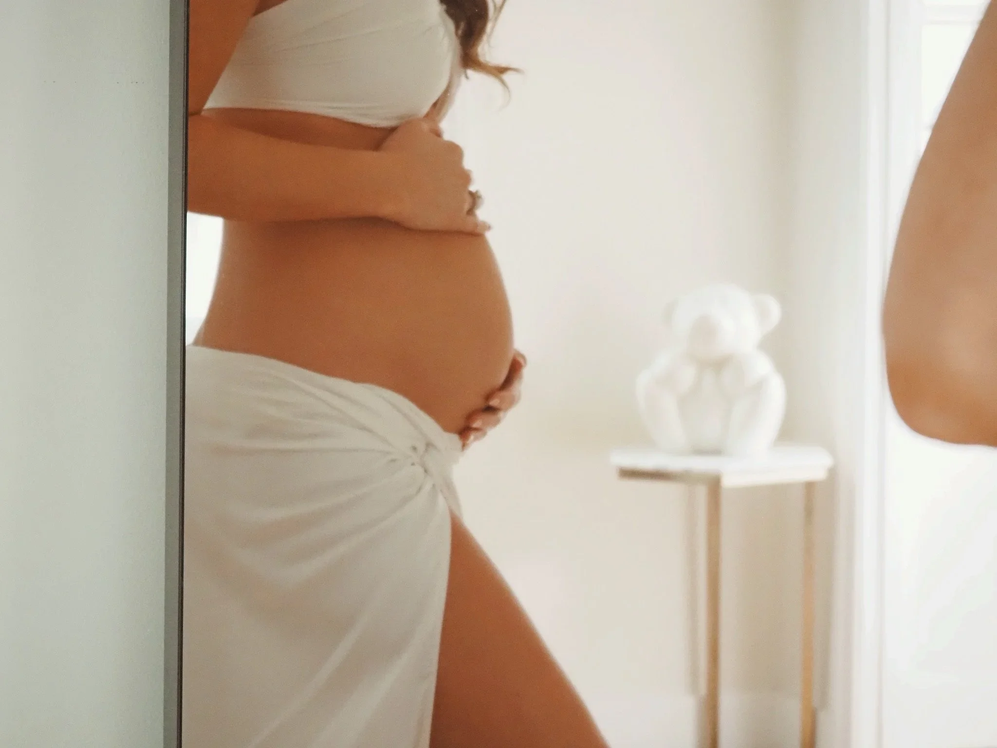 Close-up of a pregnant woman holding her belly, with her shirt lifted, standing in a softly lit room near a mirror. In the background, there is a white teddy bear on a small table.