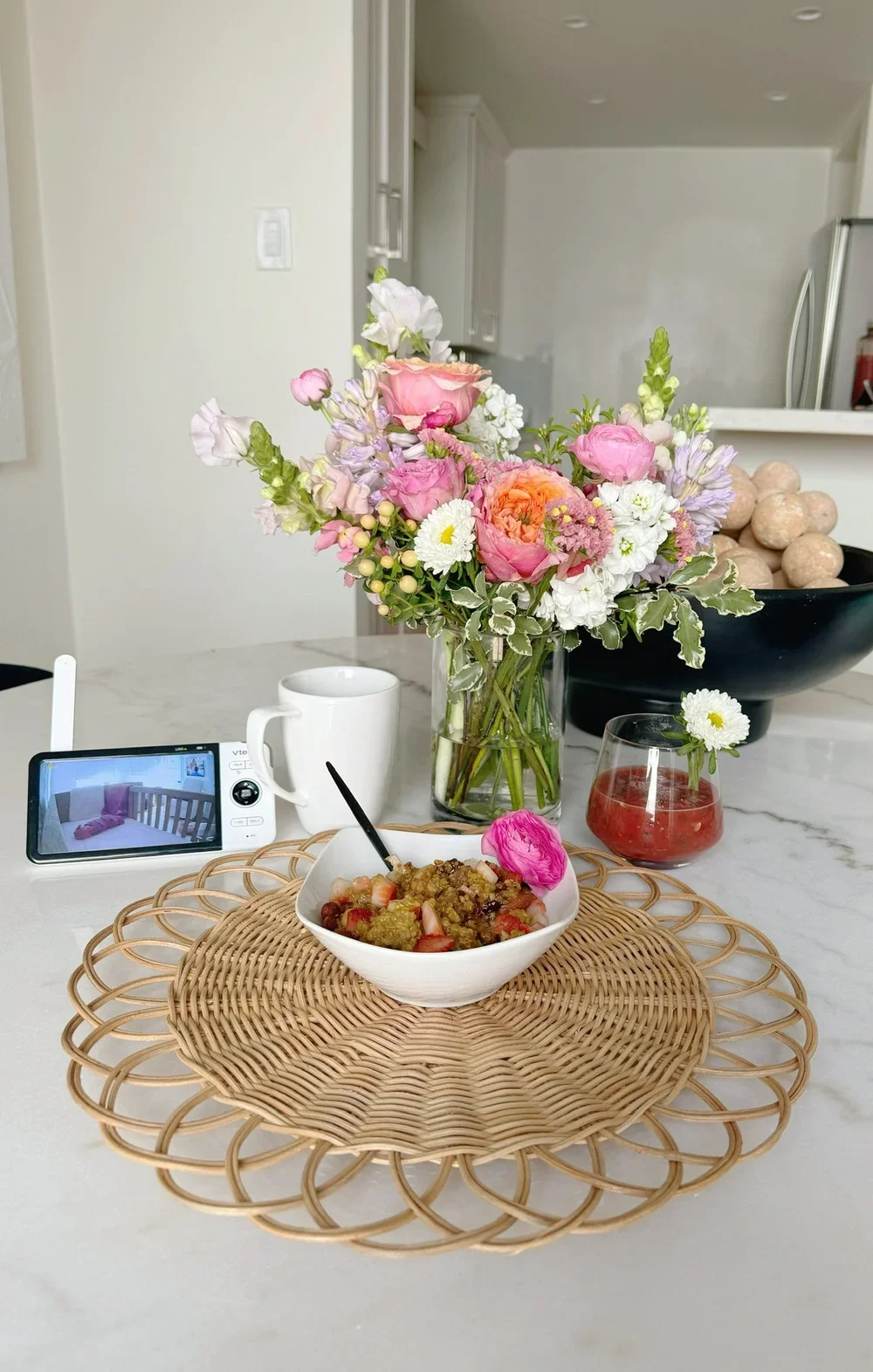 A table with a bowl of cereal, a spoon, a mug, a glass of strawberry beverage, a small daisy flower, a large vase with pink and white flowers, a black bowl with round objects, and a smartphone on a woven placemat in a kitchen.