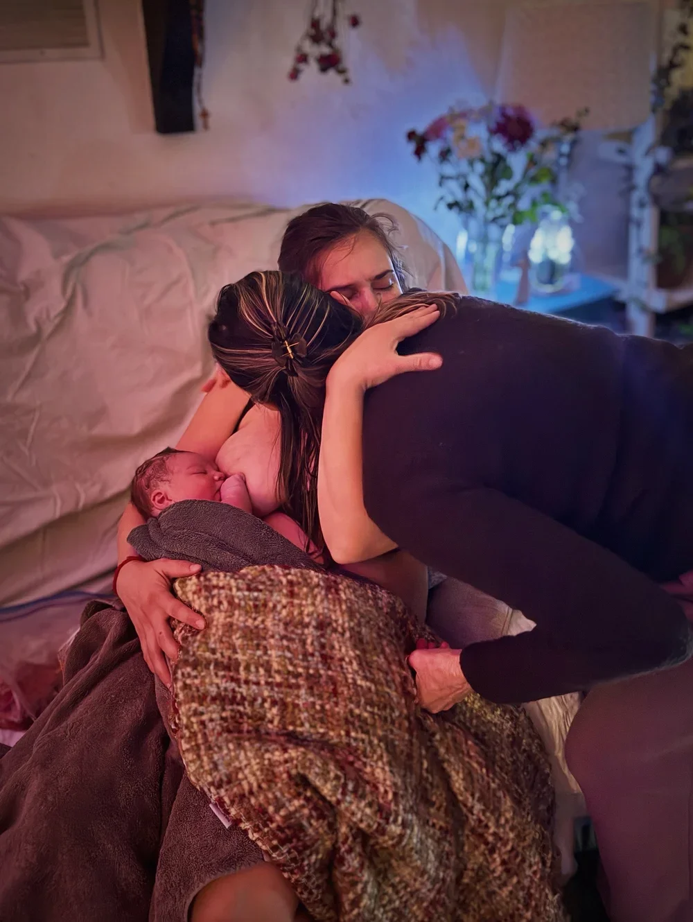 A woman holding a newborn baby in her arms while another woman hugs her, both women smiling and embracing hugs in a warmly lit room with flowers in the background.