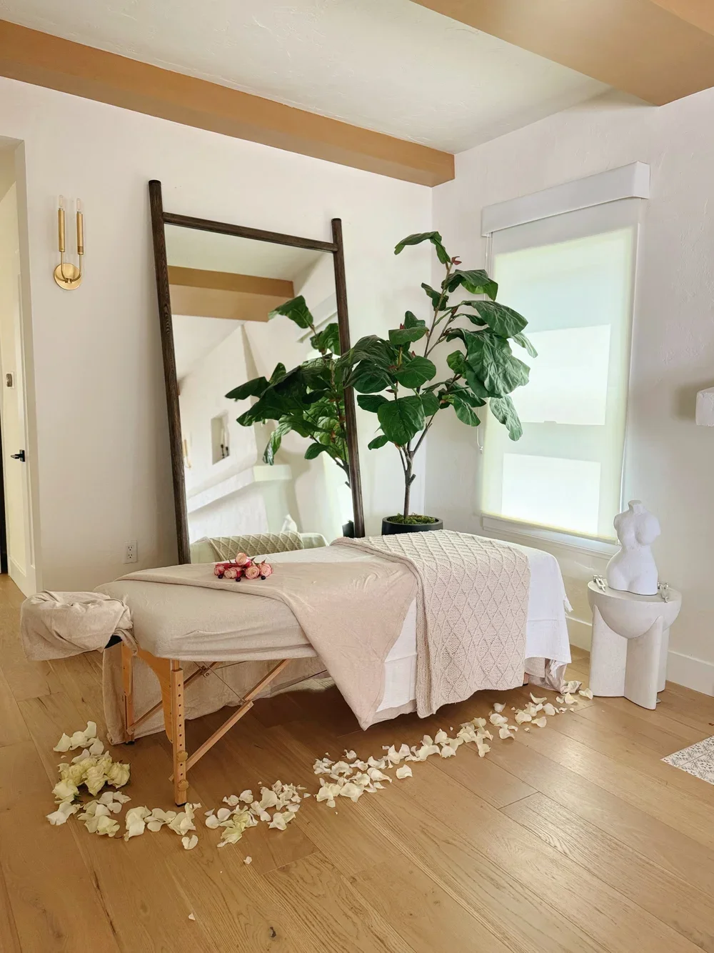 A massage table draped with white and beige towels, surrounded by flower petals on the wooden floor, with a large potted plant, a mirror, and modern decor in a bright, minimalist room.