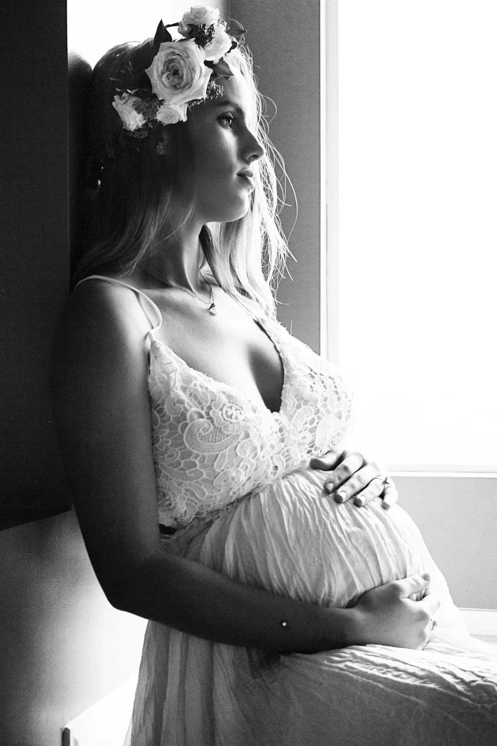 A pregnant woman wearing a lace top and a floral headband, sitting by a window, gently holding her belly, in black and white.