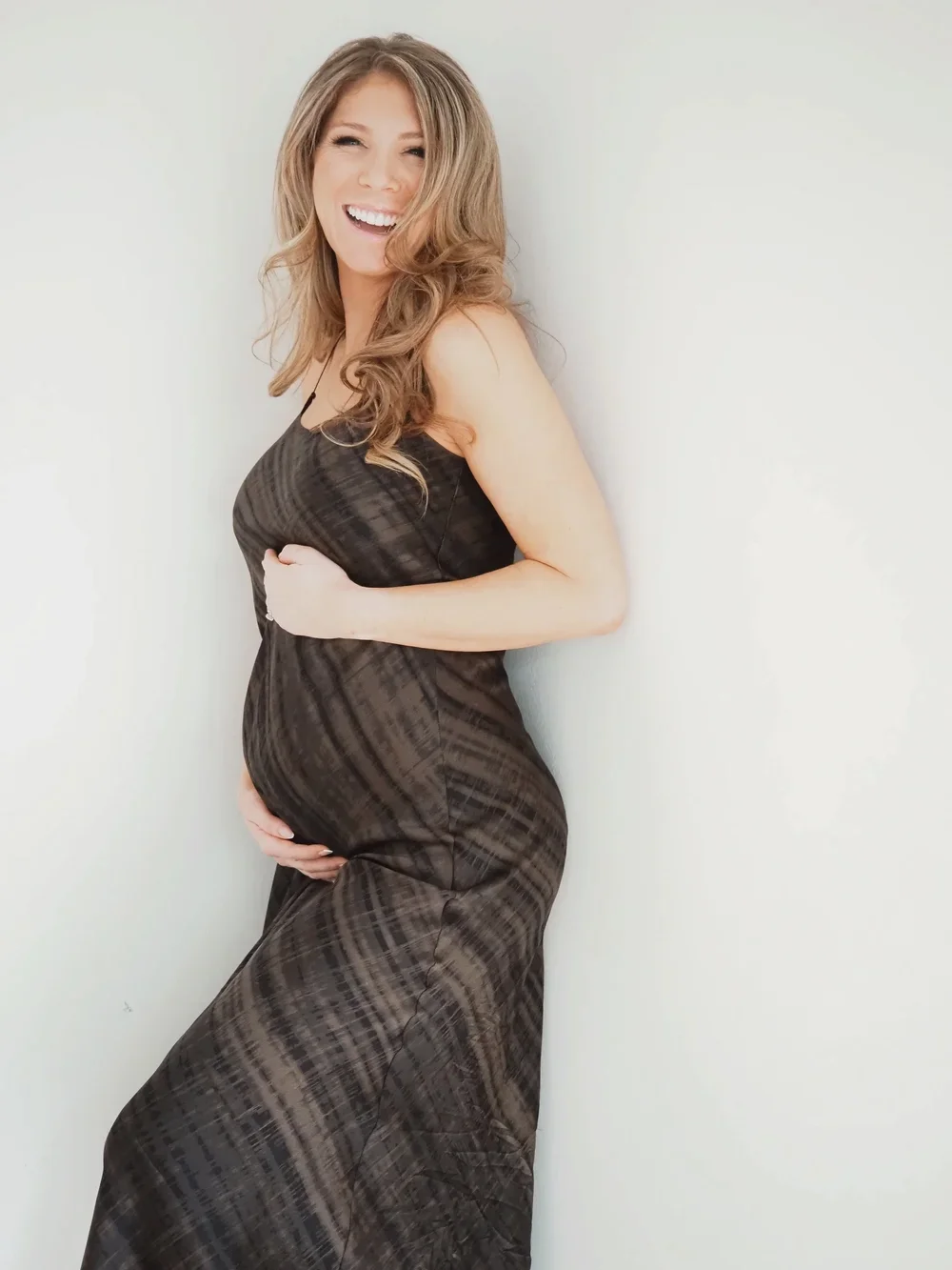 A pregnant woman with long, wavy blonde hair, smiling and standing against a plain white wall, wearing a sleeveless black dress.