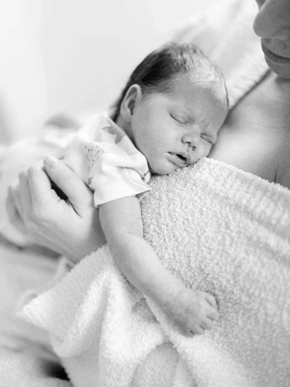 Black and white photo of a newborn baby peacefully sleeping on a fuzzy blanket, cradled in an adult's arm.