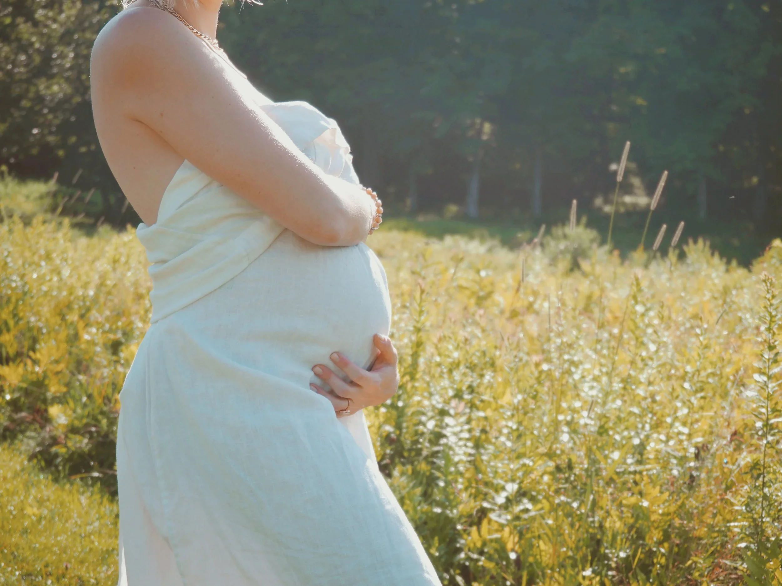 Pregnant woman standing outdoors in a field of yellow flowers, wearing a white dress, with arms crossed over her baby bump.