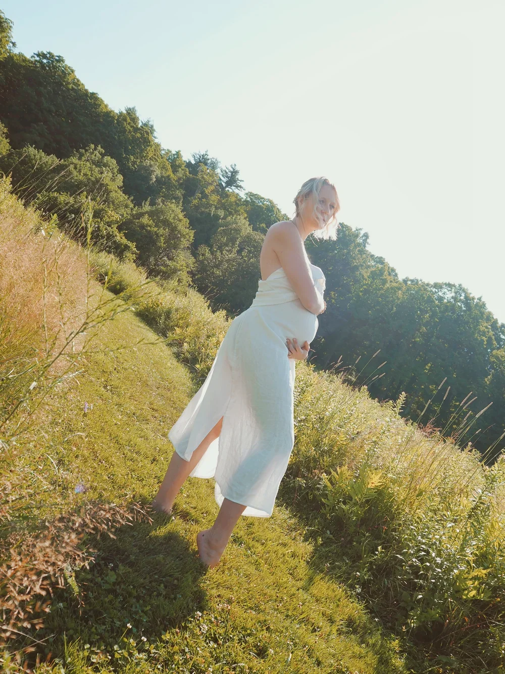 Pregnant woman in a white dress standing barefoot on grass in a sunny outdoor setting with green trees in the background.