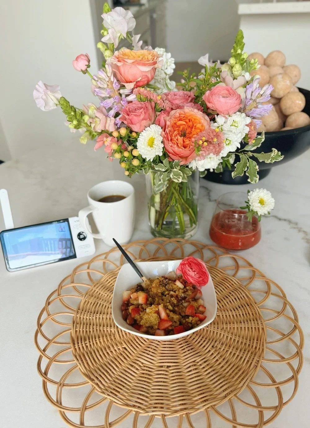 A bouquet of pink, white, and purple flowers in a glass vase on a white marble table, with a bowl of chopped fruit topped with a pink flower, a cup of coffee, a small bottle of sauce, and a small white flower in a glass