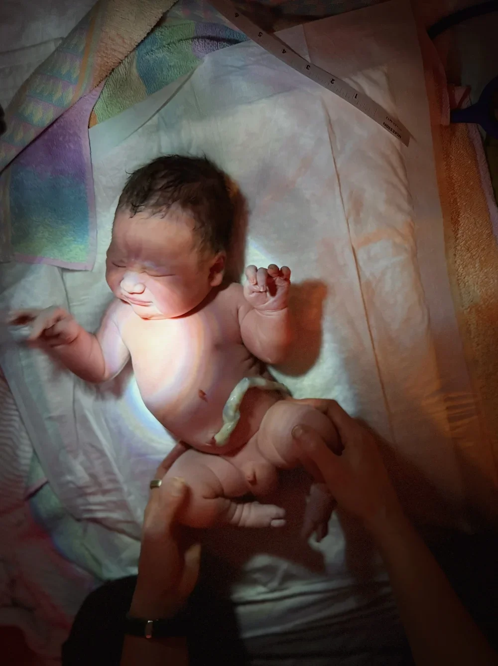 Newborn baby lying on a white cloth during birth, with emergency medical lighting, showing the umbilical cord.