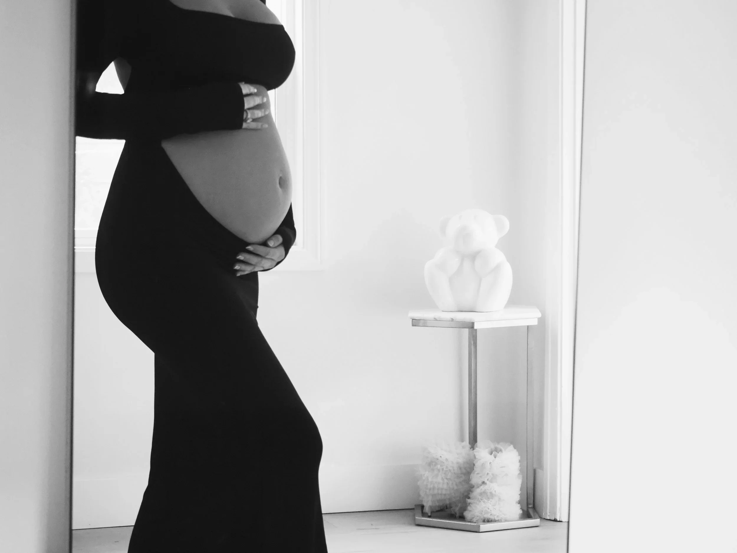 A pregnant woman wearing a black dress standing sideways, cradling her belly, in a bright room near a window with a white bear sculpture on a small stand, and fluffy decorations on the floor.