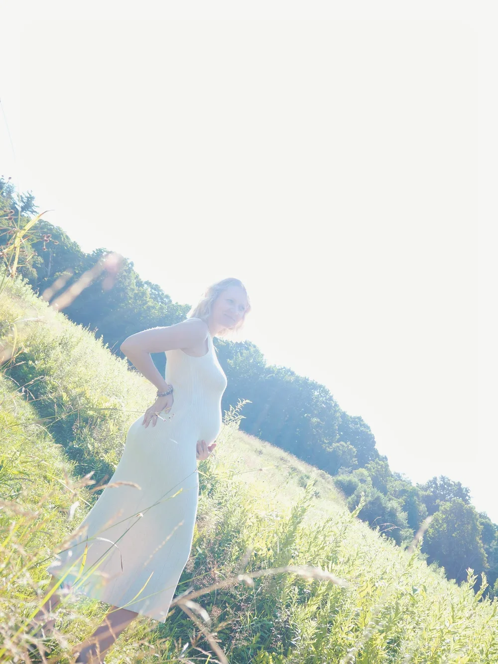 A pregnant woman wearing a white dress standing in a sunlit grassy field, with trees and a hill in the background.