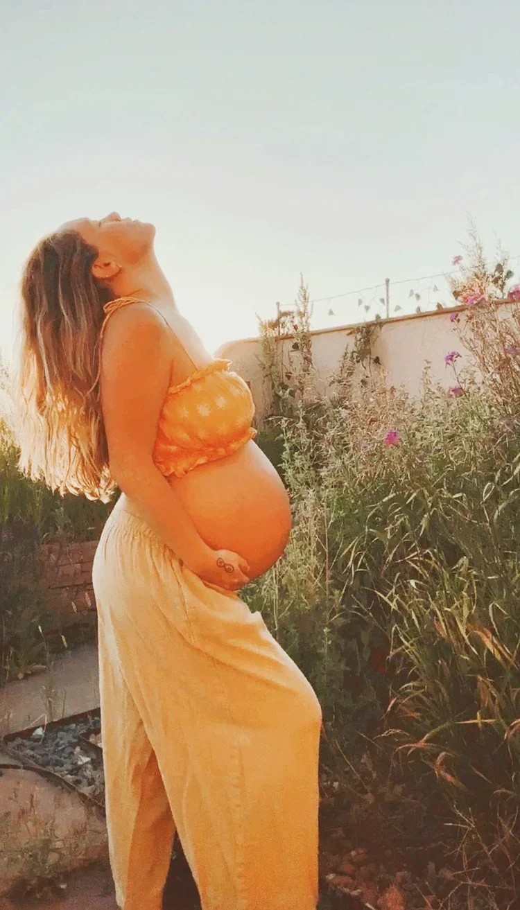 A pregnant woman standing outdoors at sunset, gently holding her baby bump and smiling with her eyes closed.