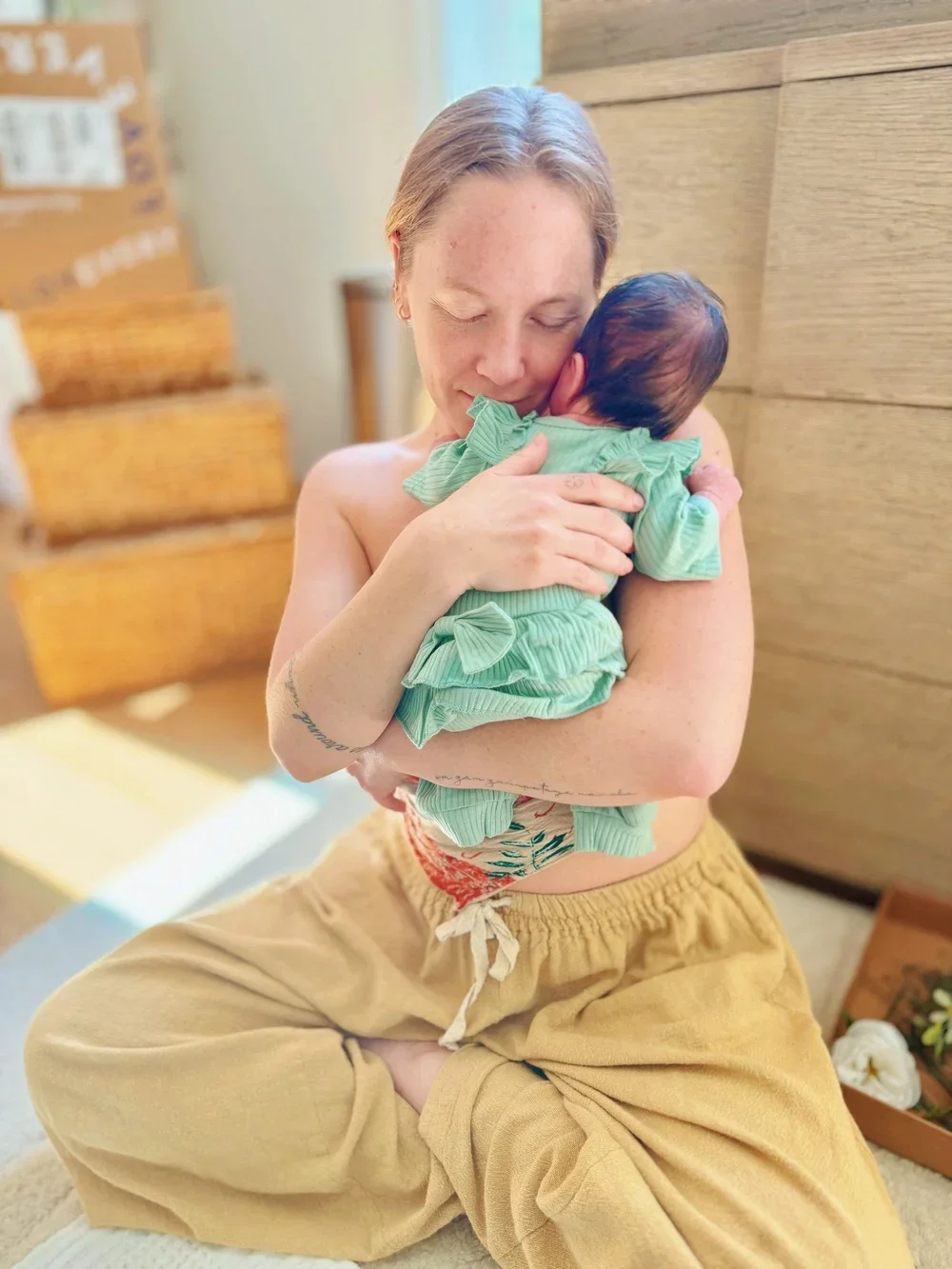 A woman postpartum doula, sitting cross-legged, holding and cuddling a newborn baby dressed in a green outfit. She has her eyes closed, smiling gently, and is seated indoors with a wooden wall and stacked cardboard boxes in the background.