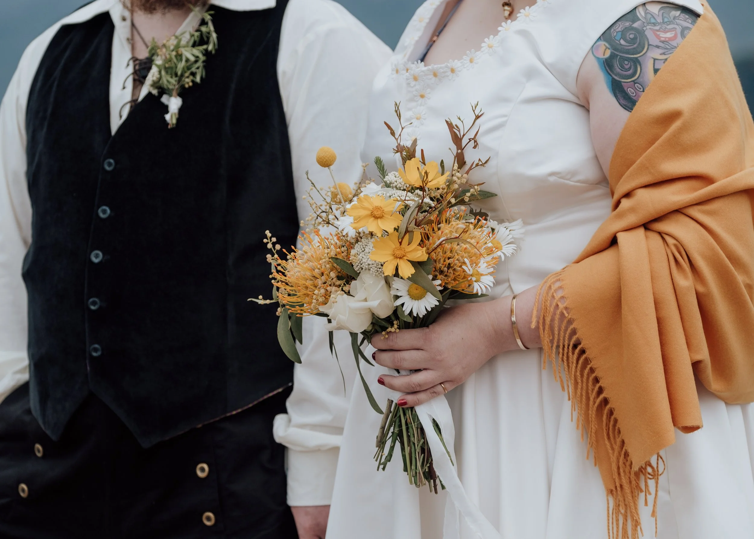 A close-up of a bride holding a bouquet of yellow, white, and orange flowers beside her groom — bright, elegant, and full of warmth.