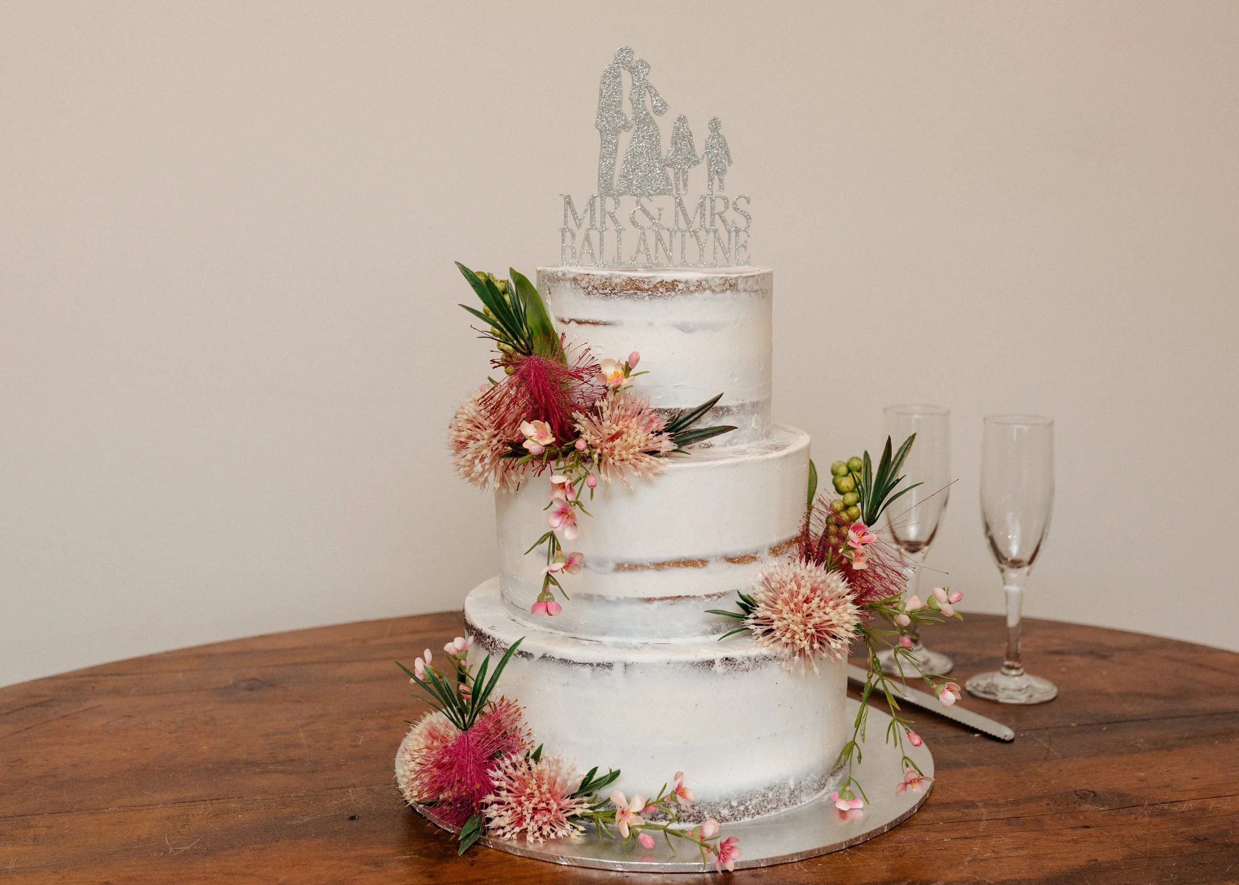 Three-tiered white wedding cake decorated with pink and green flowers and foliage, topped with a glittery cake topper that says "Mr & Mrs Ballantyne" with silhouette figures of a woman and children on top, placed on a wooden table with two champagne glasses in the background.