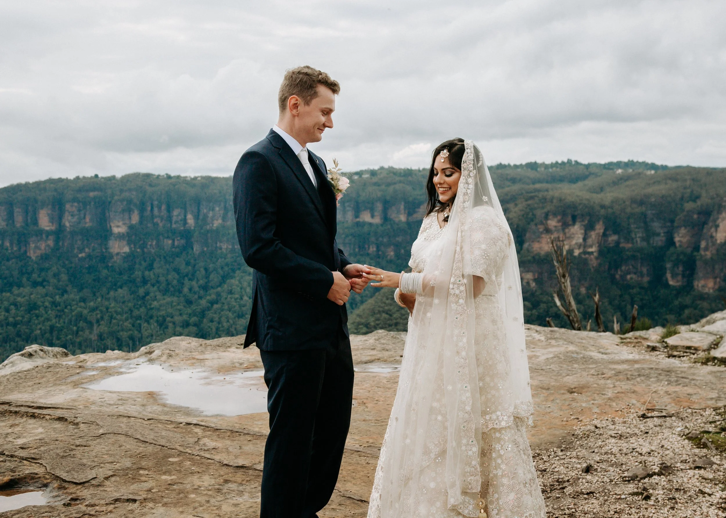 A bride and groom standing outdoors on a rocky area, exchanging vows with a mountainous landscape in the background. Blue Mountains elopement vows scenic mountain wedding Penrith intimate ceremony
