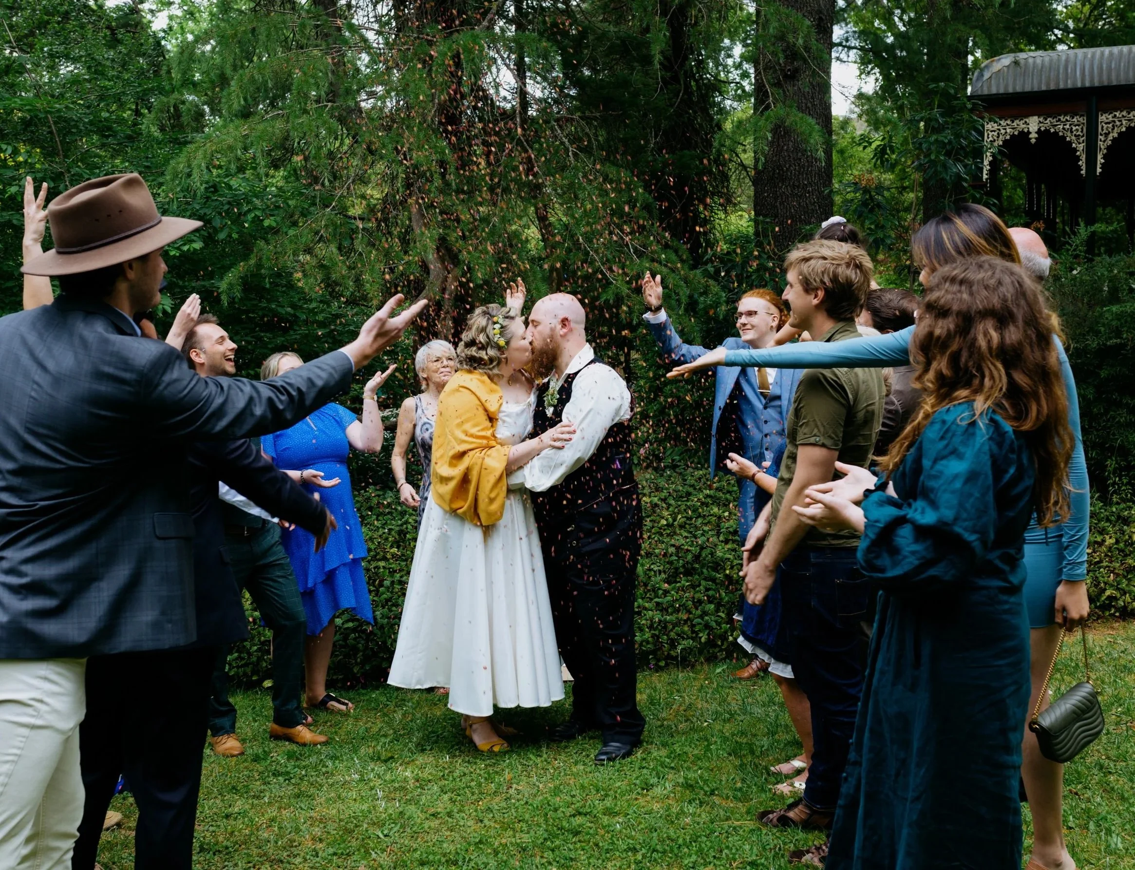 A wedding celebration outdoors with a couple kissing in the center, surrounded by guests throwing flower petals.