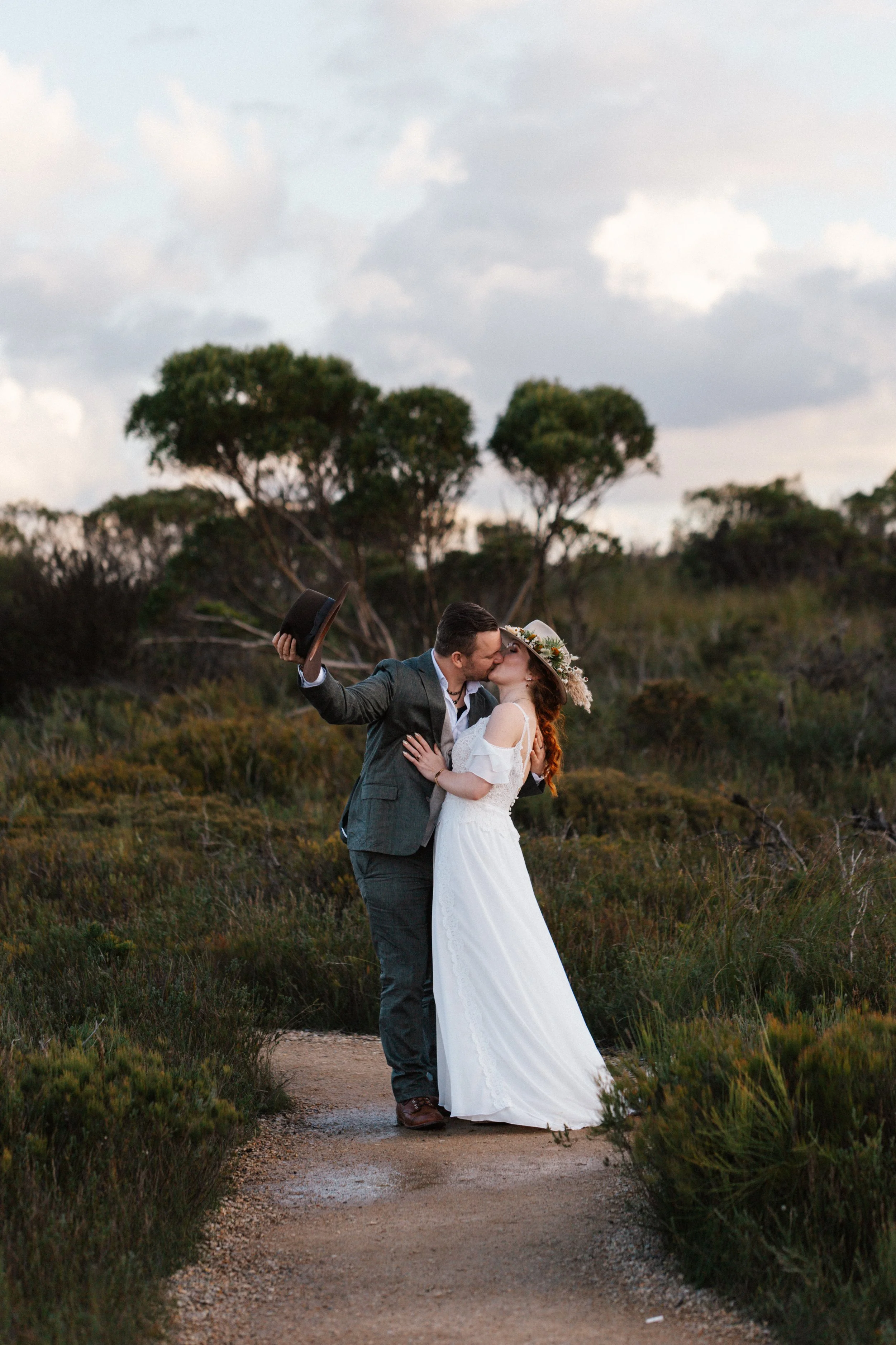 A couple kissing outdoors on a dirt path surrounded by greenery, the woman wearing a white dress and floral hat, the man in a gray suit, holding a hat in his hand.