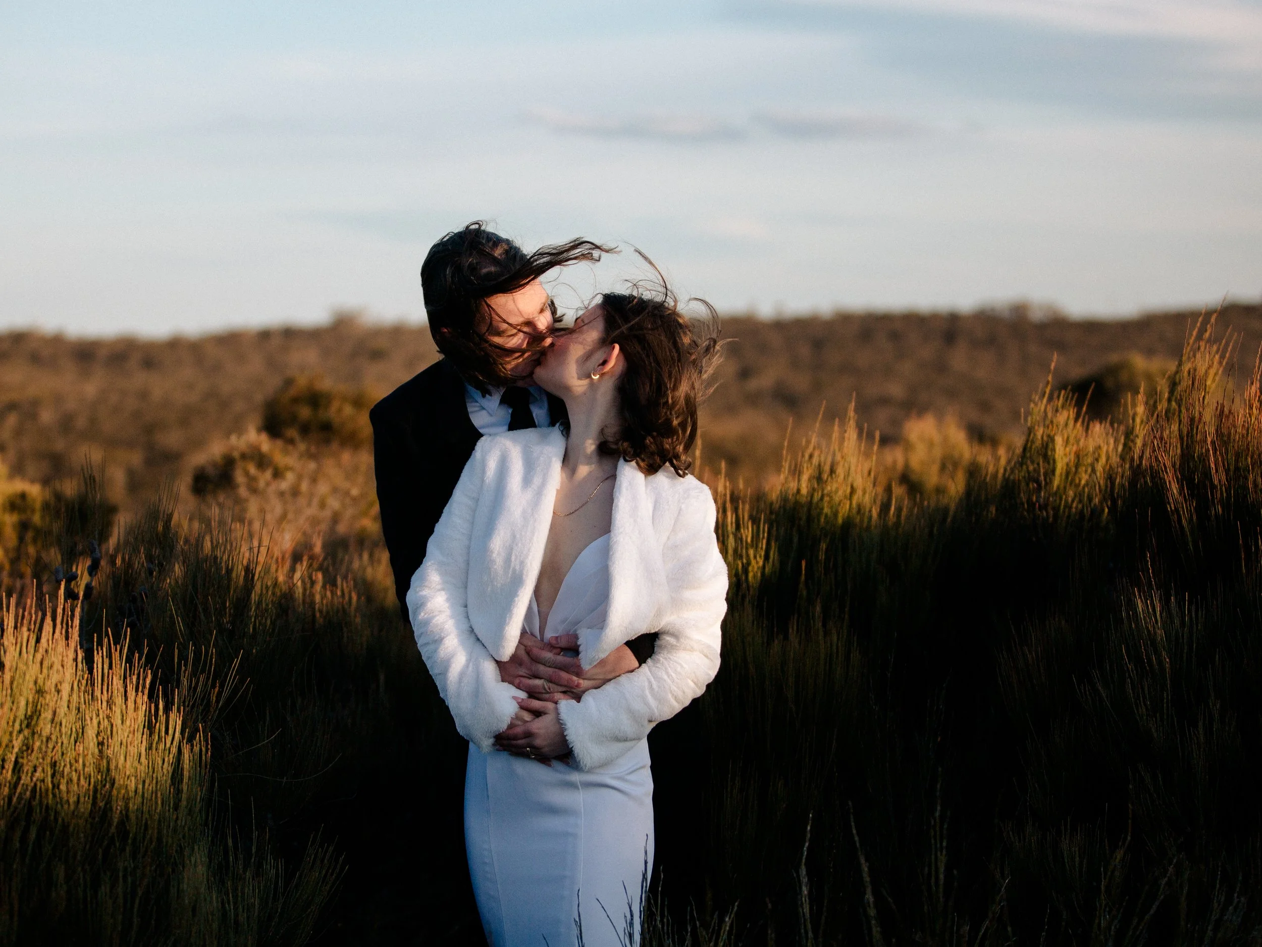 A couple sharing a kiss outdoors in a field at sunset, with the wind blowing through their hair.
