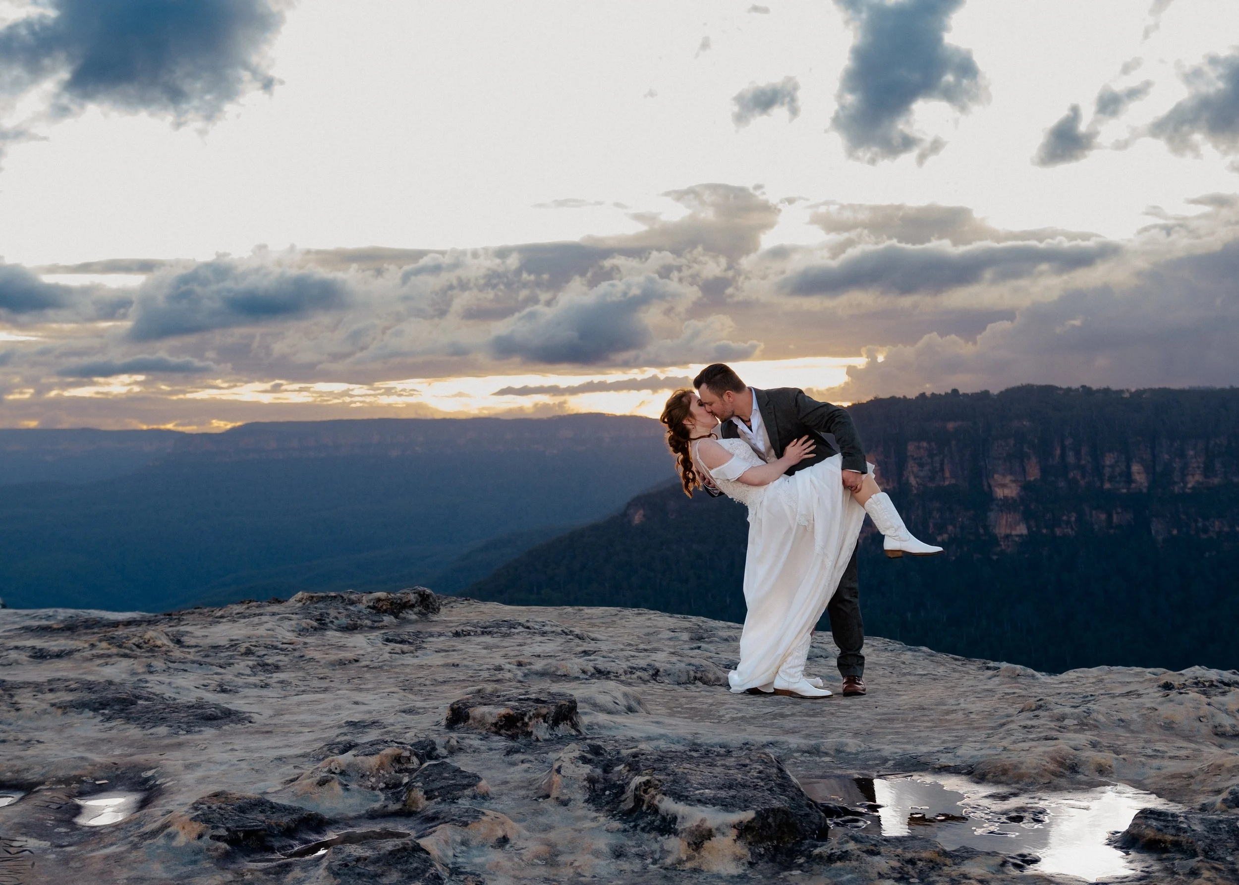 A newlywed couple sharing a kiss on a rocky ledge during sunset, with mountains and a cloudy sky in the background.