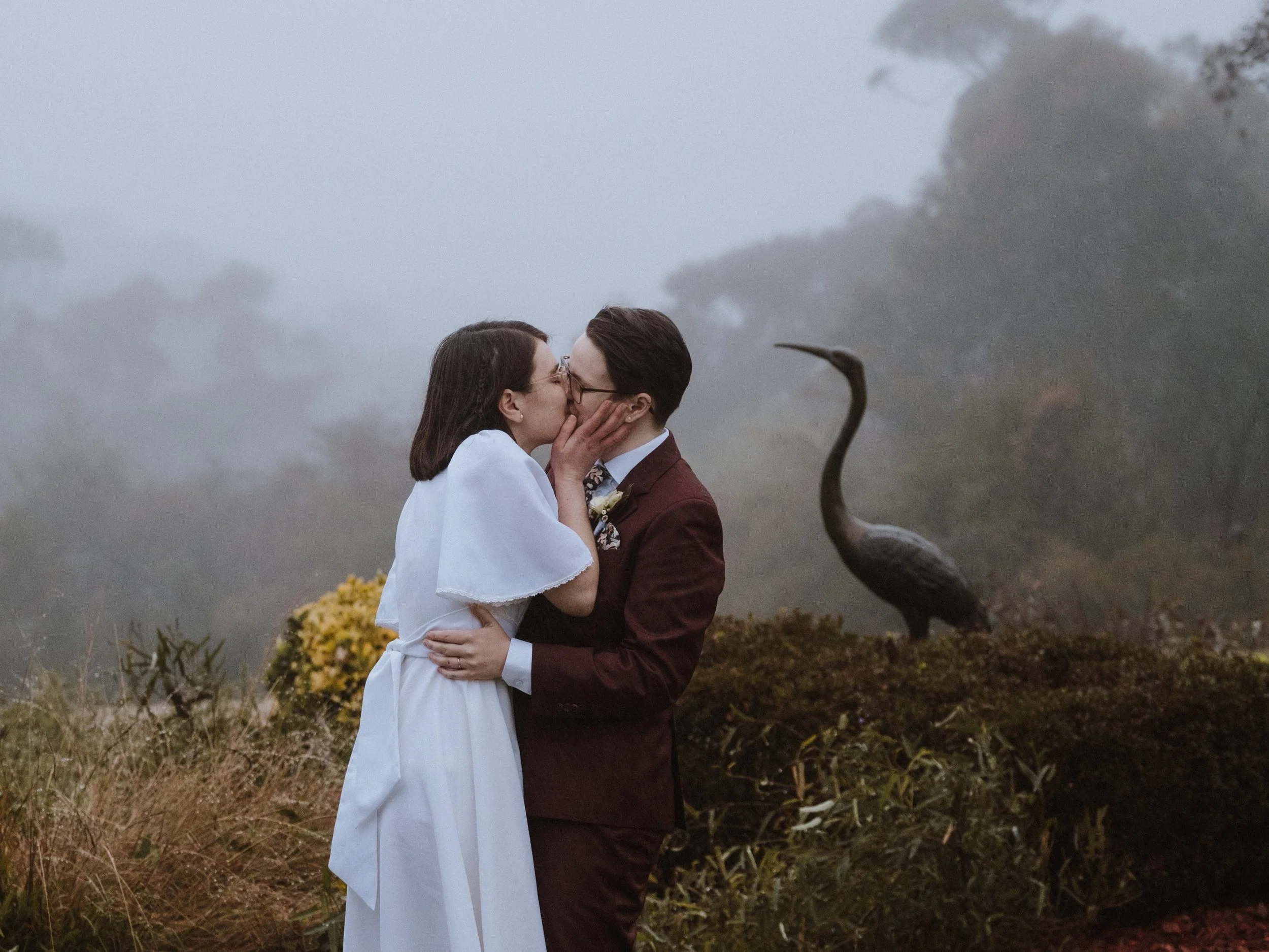 A couple kissing outdoors in front of a bronze heron statue with foggy trees in the background. Blue Mountains elopement couple romantic outdoor wedding Penrith boutique photography.