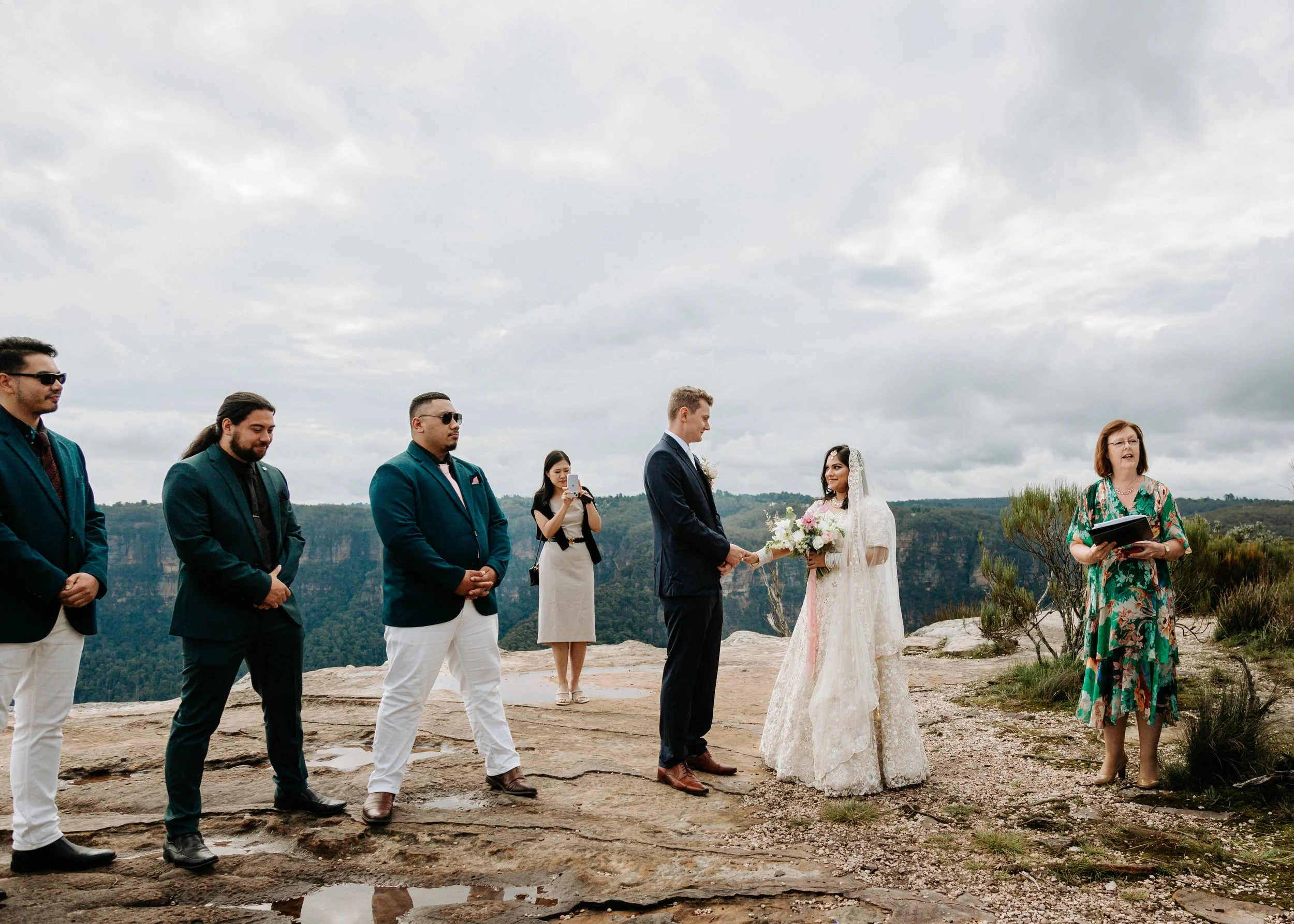 A couple gets married outdoors on a rocky landscape with a cloudy sky, exchanging vows while friends and a officiant look on. Blue Mountains outdoor elopement ceremony boutique wedding Penrith intimate vows