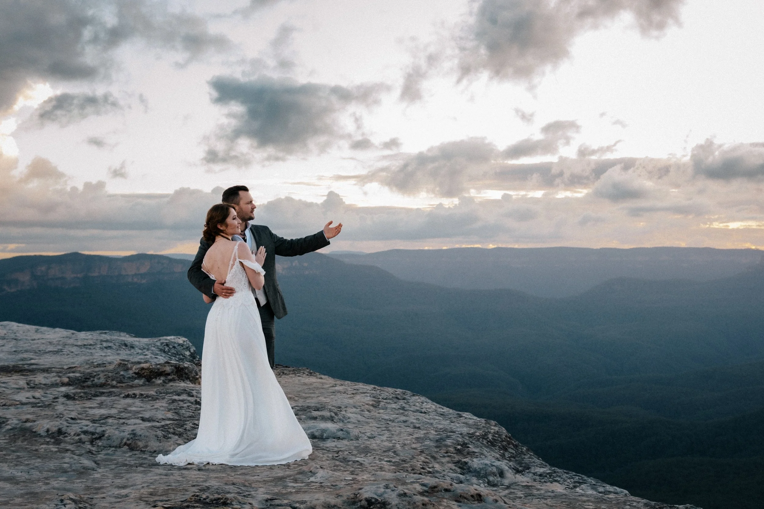 A newlywed couple stands on a cliff edge at sunset, surrounded by soft mountain light — the perfect moment for a romantic Blue Mountains elopement.