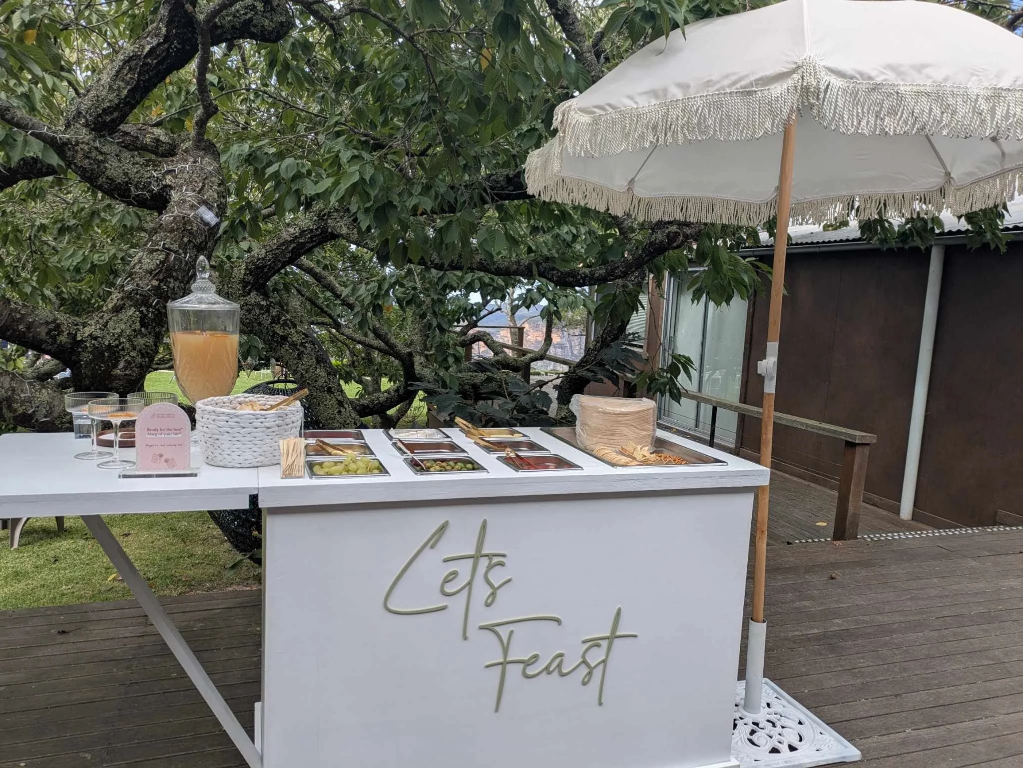 Outdoor brunch station with a white table under a white parasol, featuring beverages, snacks, and small food containers, set on a wooden deck with a large leafy tree in the background.
