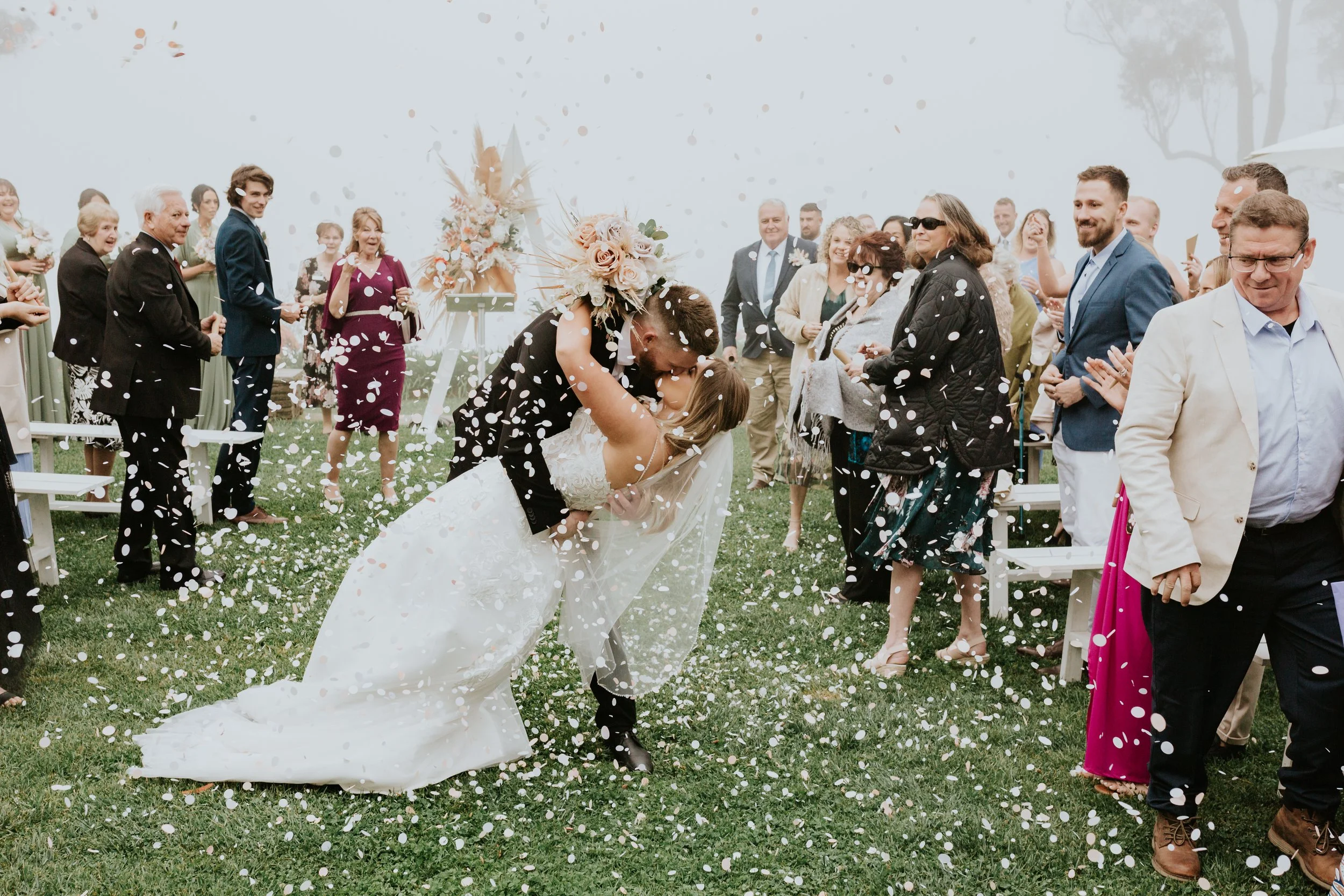 Couple dancing at their wedding with guests around, confetti falling, outdoor setting with foggy sky.