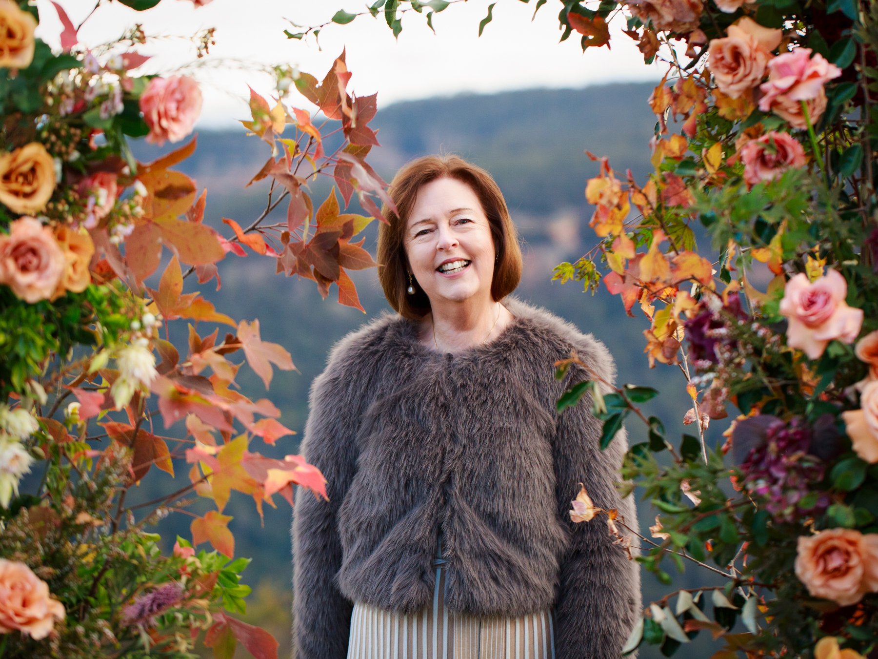 A woman in a gray fur jacket smiling in a garden with pink and peach flowers and autumn leaves, with hills in the background.