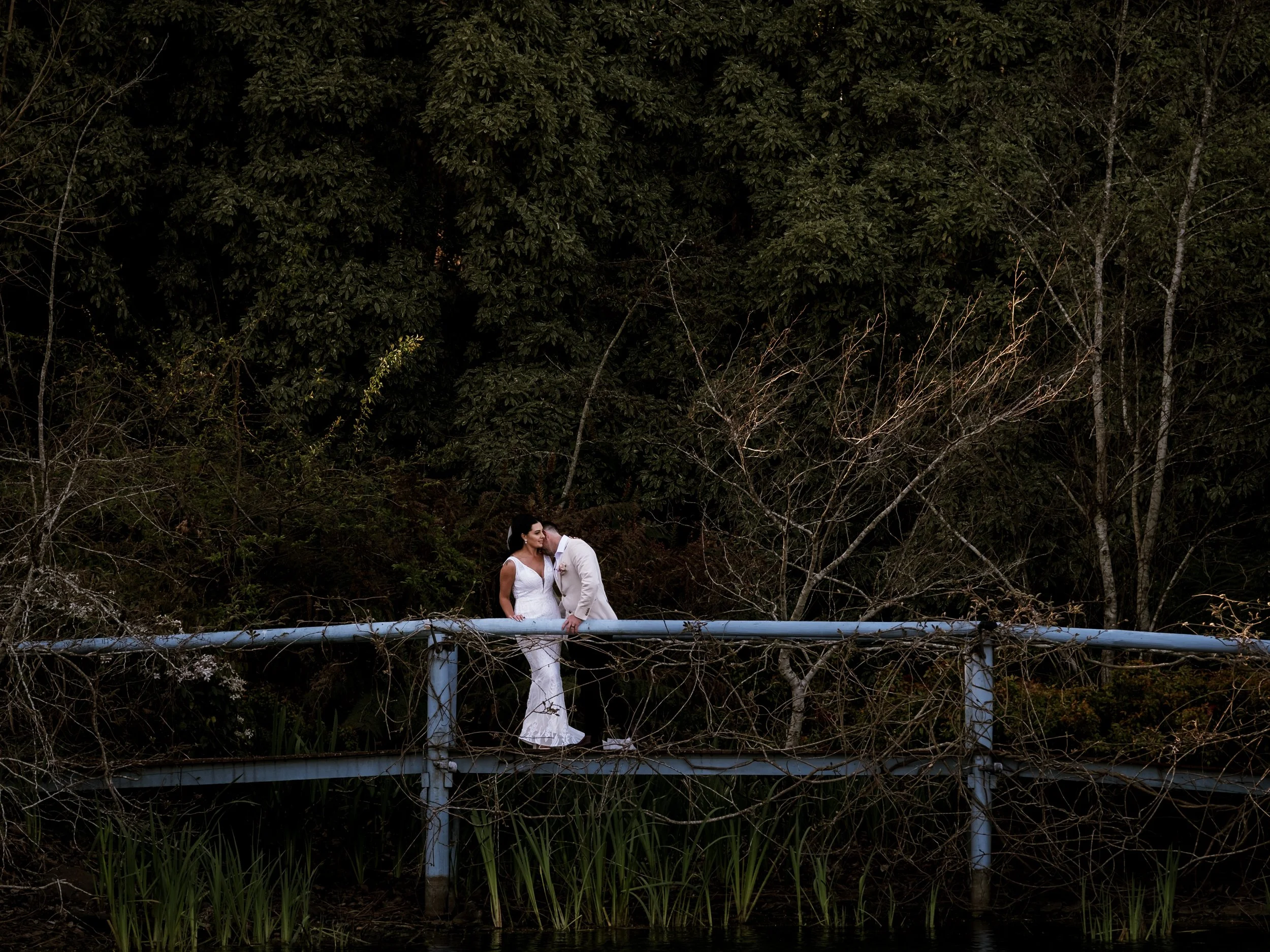 A bride and groom standing on a weathered blue bridge surrounded by dense trees and bushes, sharing a moment of affection.