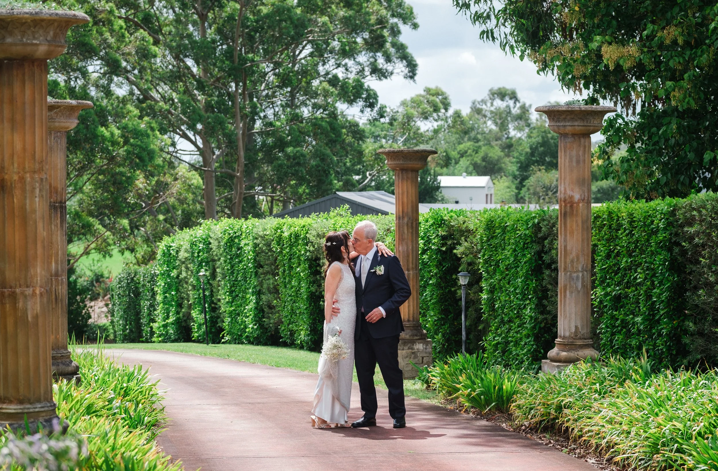 A bride and groom sharing a kiss on a garden pathway lined with green hedges and old columns, with trees and a house in the background.