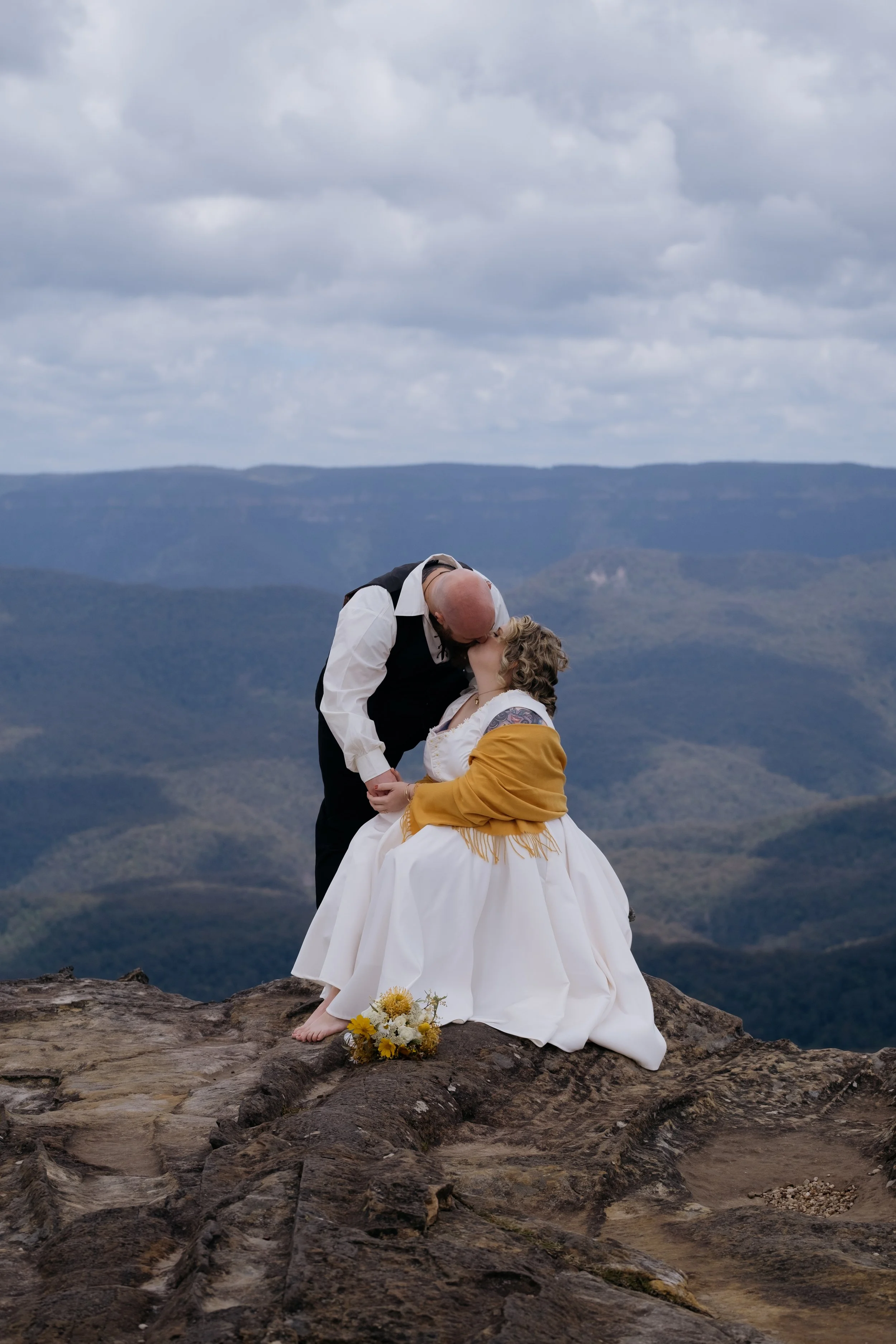 A couple dressed in vintage wedding attire sharing a kiss on a rocky mountain ledge against a backdrop of clouds and mountains. Blue Mountains vintage elopement photography romantic mountain wedding Penrith