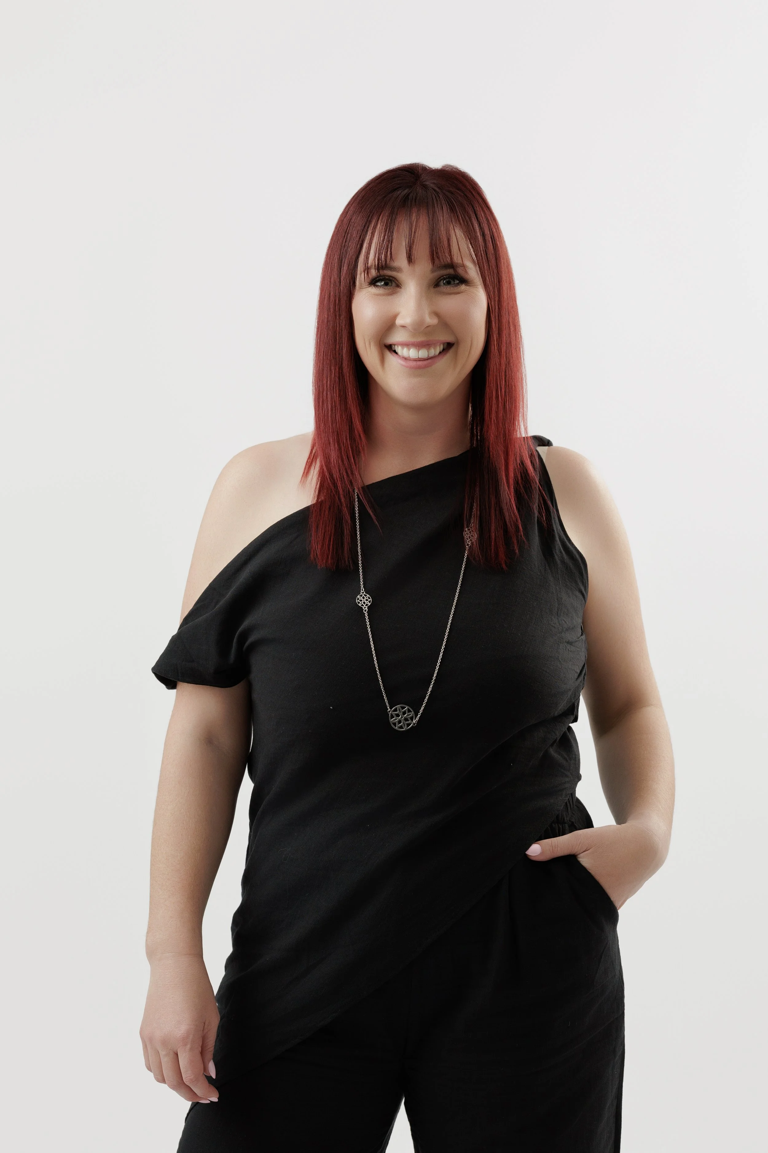 A woman with red hair wearing a black, off-shoulder top and necklace, smiling with one hand in her pocket against a white background.