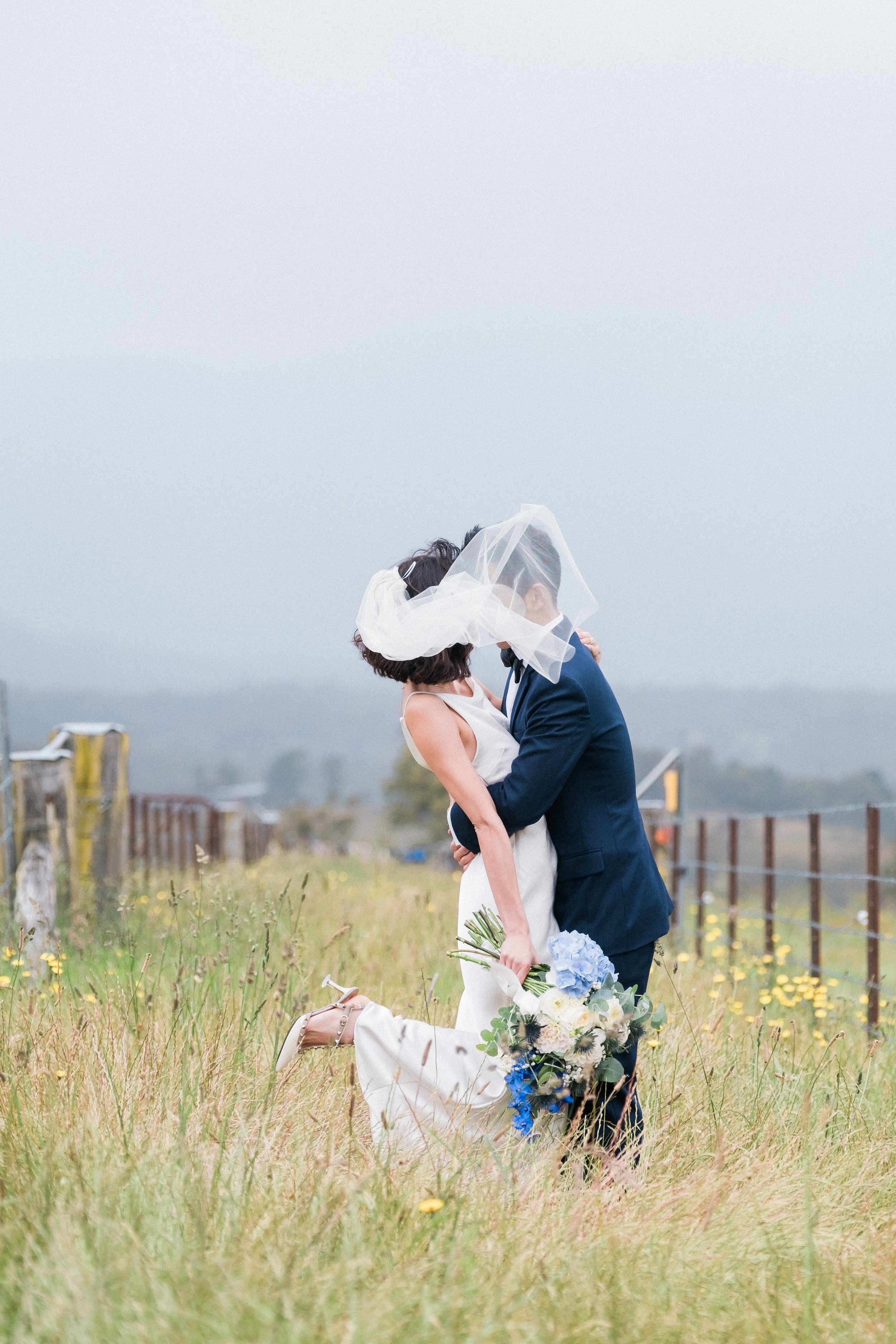 A couple in wedding attire sharing a kiss outdoors, with the bride holding a bouquet of white and blue flowers, and the groom lifting the bride.