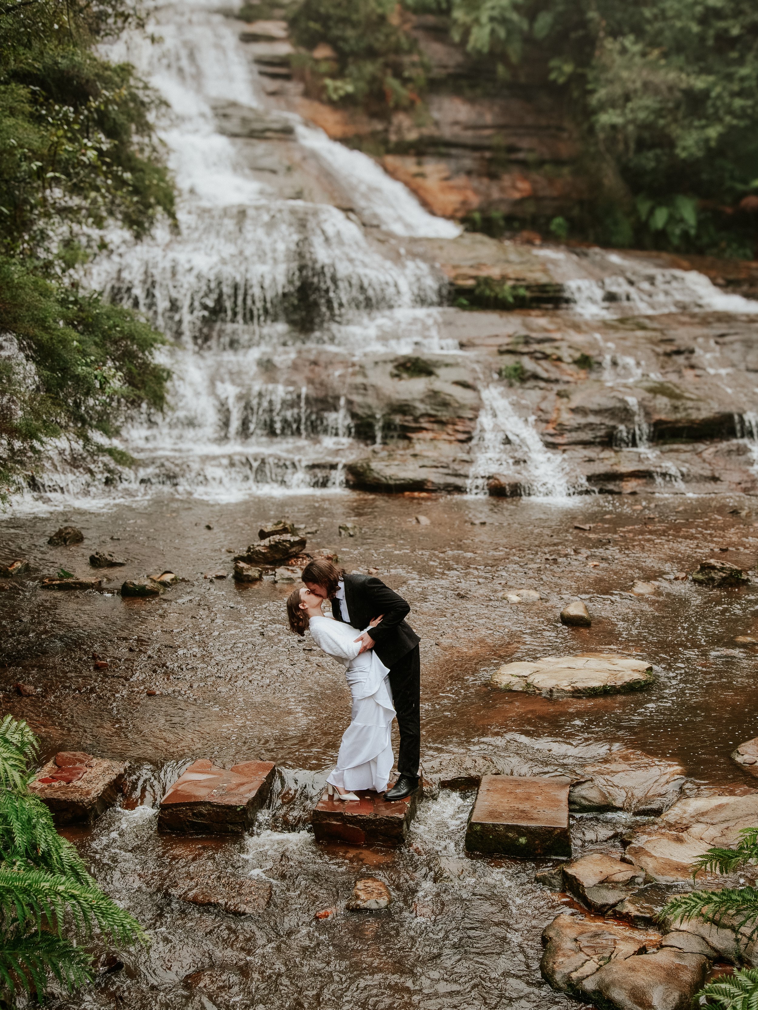 A couple dressed in wedding attire sharing a kiss on stepping stones in a shallow creek with a waterfall in the background.