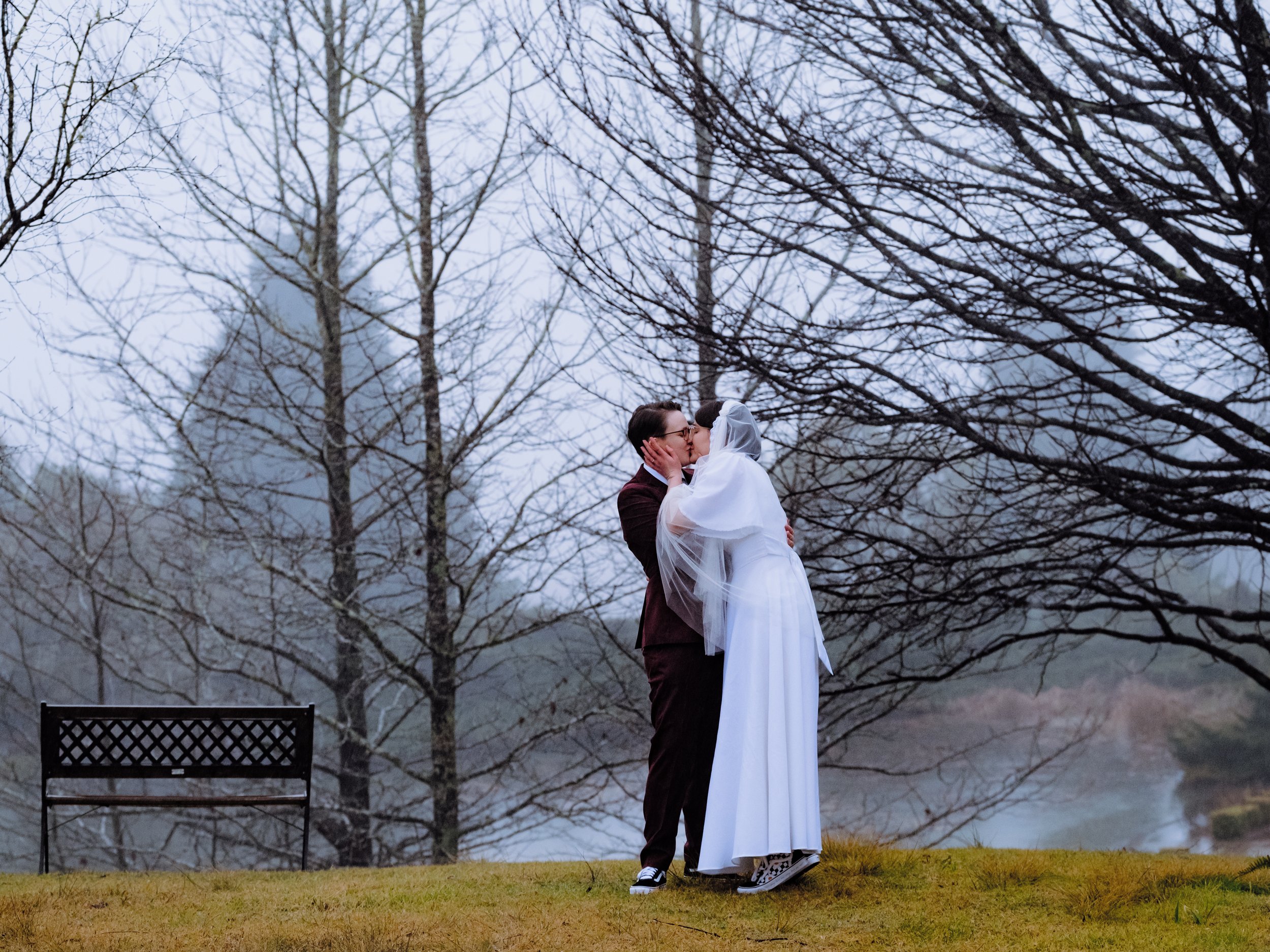 A couple is sharing a kiss outdoors on a foggy day, with trees around and a bench in the background. Blue Mountains elopement couple romantic foggy wedding Penrith intimate photography
