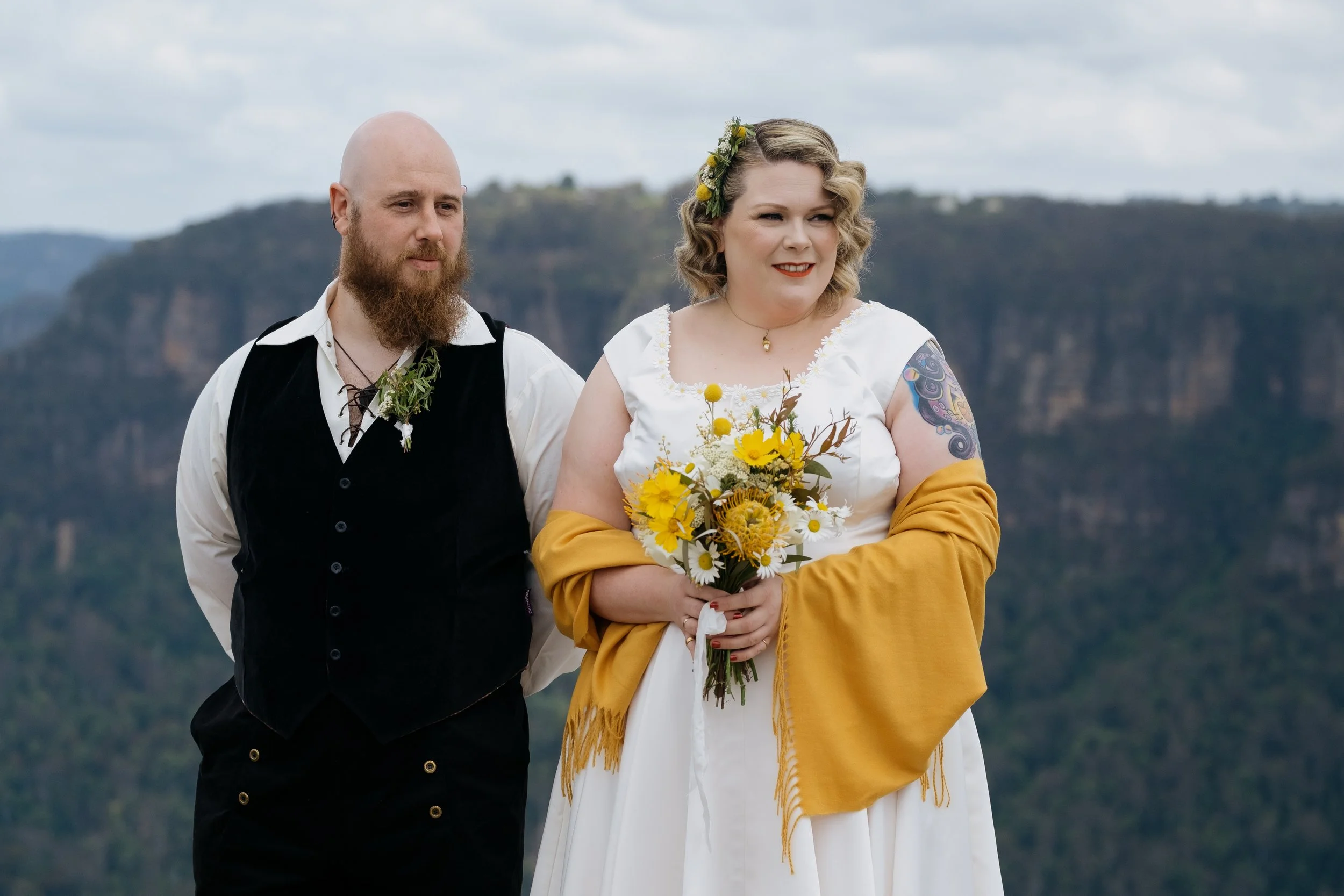 A joyful couple celebrates their mountain elopement, holding bright yellow blooms against the stunning Blue Mountains landscape.