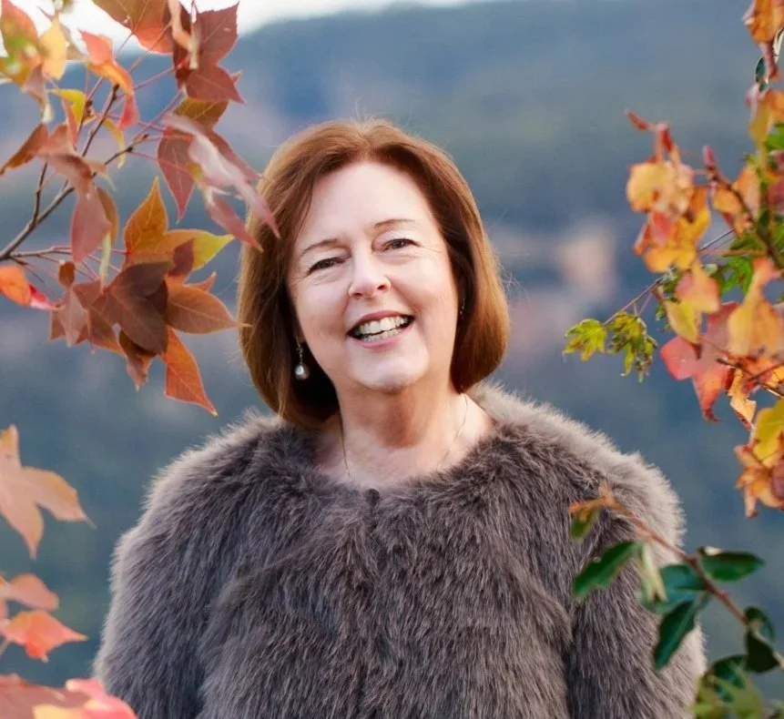 Blue Mountains wedding celebrant Karen Brown, smiling outdoors surrounded by autumn leaves and mountain scenery, wearing a soft faux fur coat.