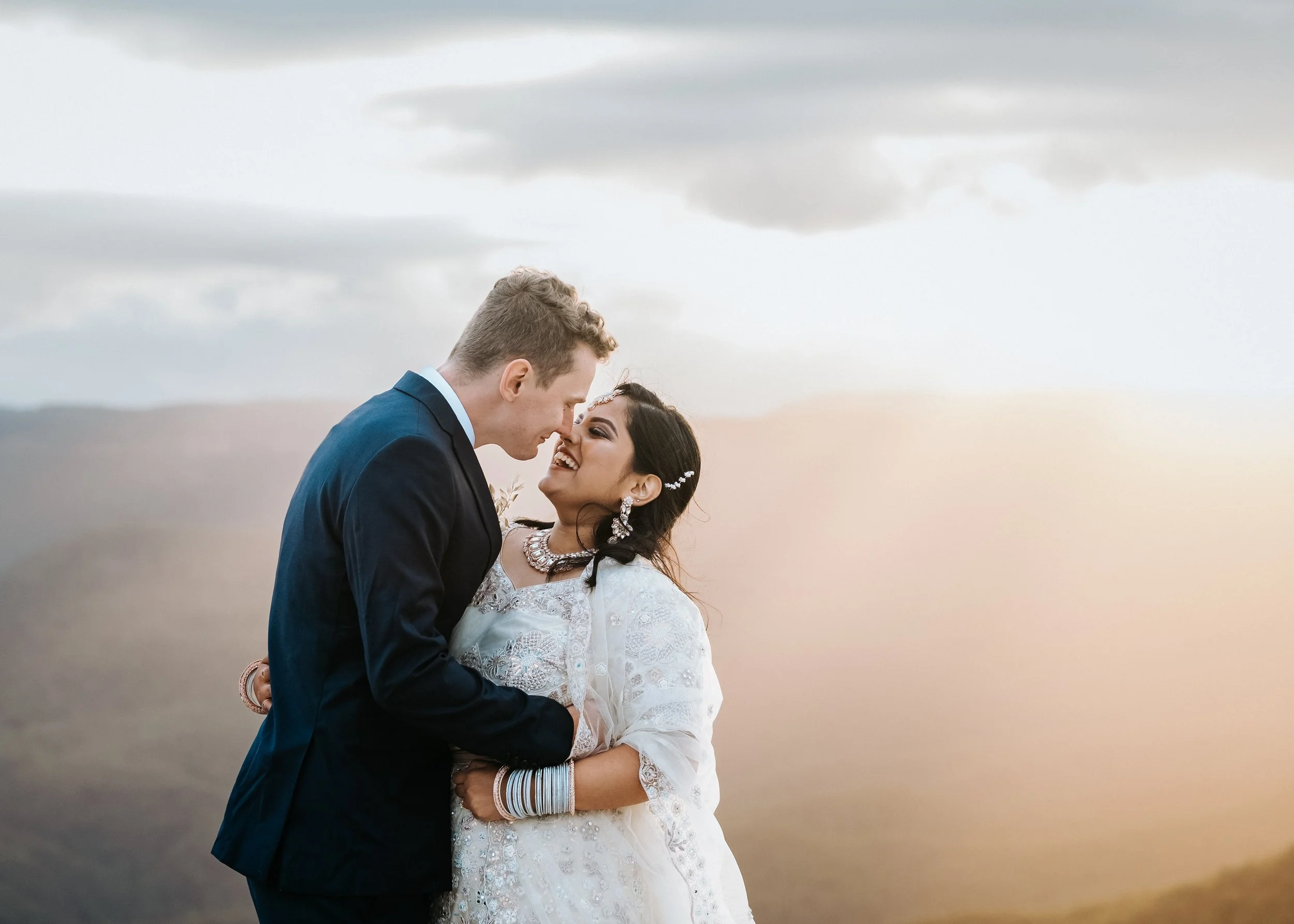 A happy couple in wedding attire smiling and embracing outdoors during sunset, with a cloudy sky in the background.