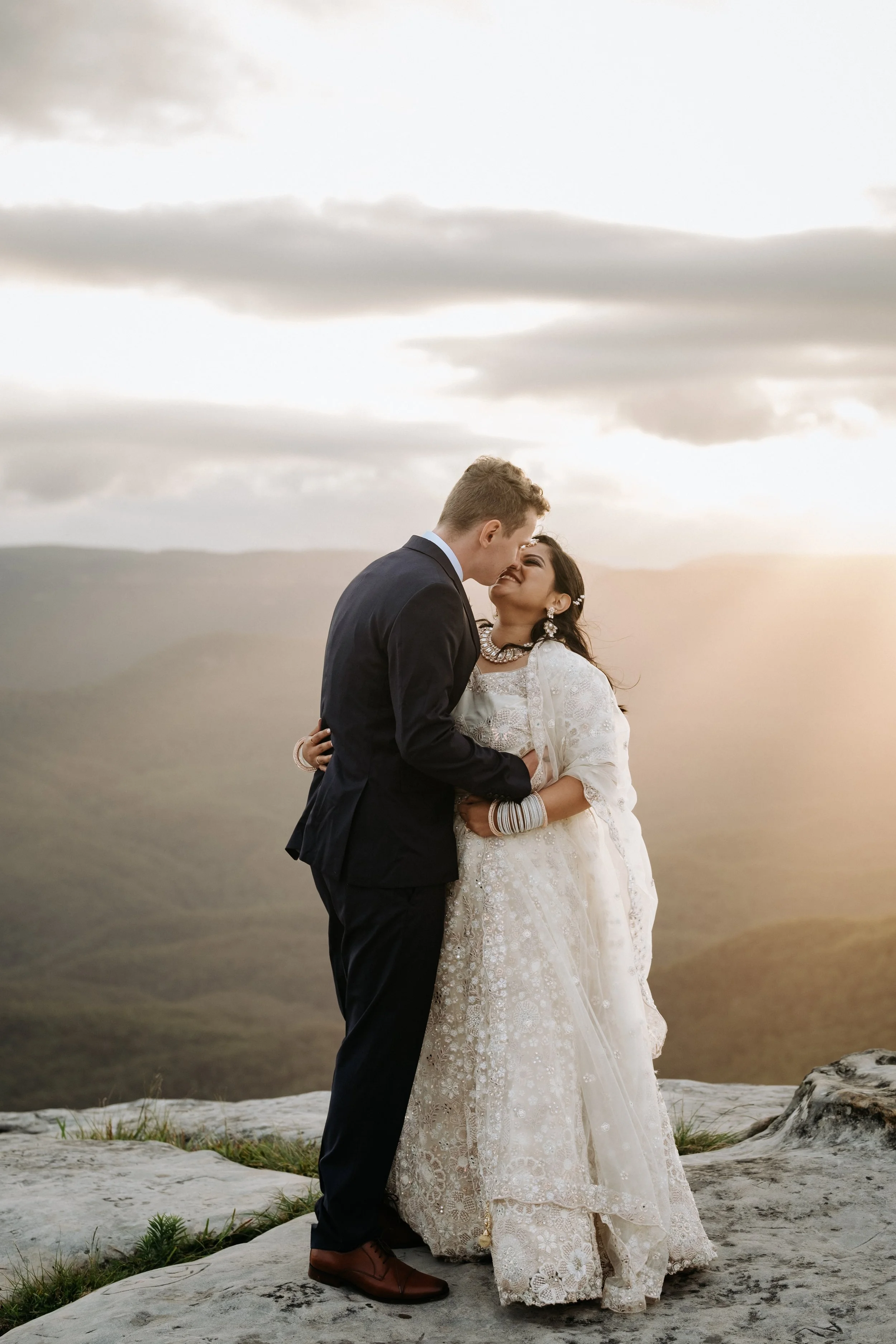 A newlywed couple sharing a kiss on a mountain during sunset, with rolling hills and dramatic clouds in the background.